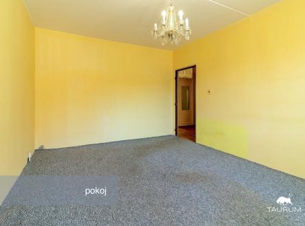 Světlý pokoj s koberci a lustr na stropě, ideální pro snadnou rekonstrukci. Vstup do další místnosti s parketovou podlahou.
