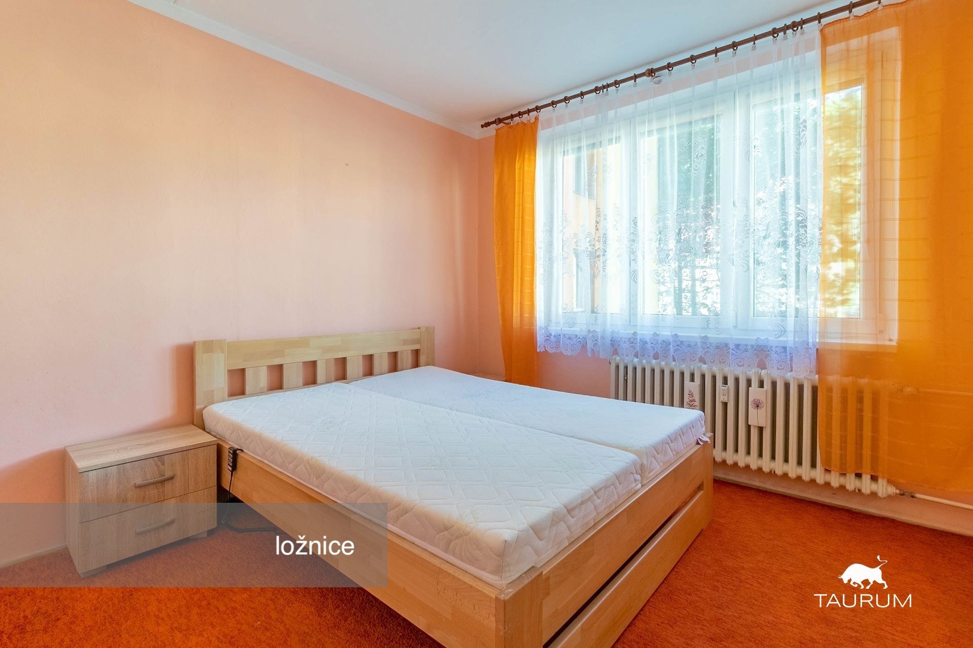 Světlá ložnice s dřevěnou postelí a velkým oknem, oranžové stěny a koberce, moderní vybavení, příjemná atmosféra a dobré osvětlení.