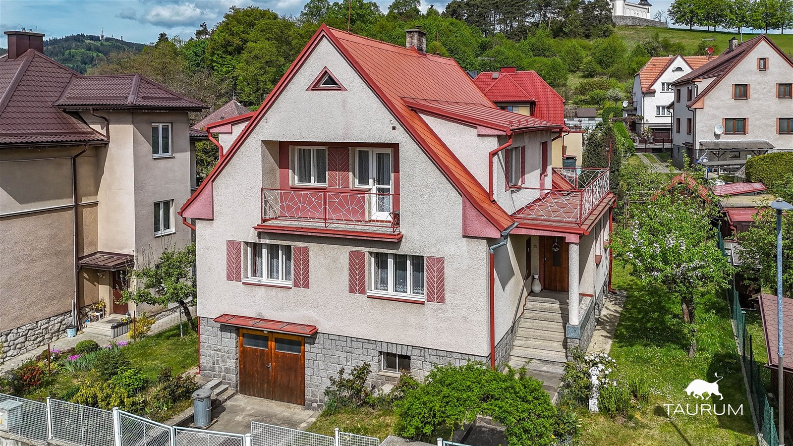 Rodinný dům s červenou sedlovou střechou, obklopený zahradou. Fasáda v kombinaci světlé omítky a kamenného soklu, prostorný balkon, klidná lokalita.