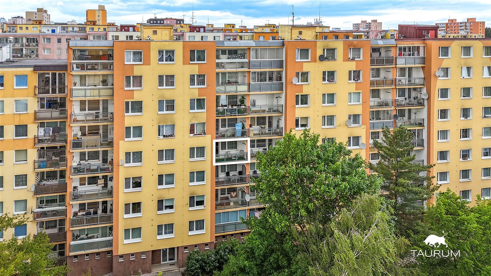 Panelový bytový dům se světlou fasádou, velká okna a balkony, vhodný pro rodiny, umístěný na sídlišti s městským výhledem