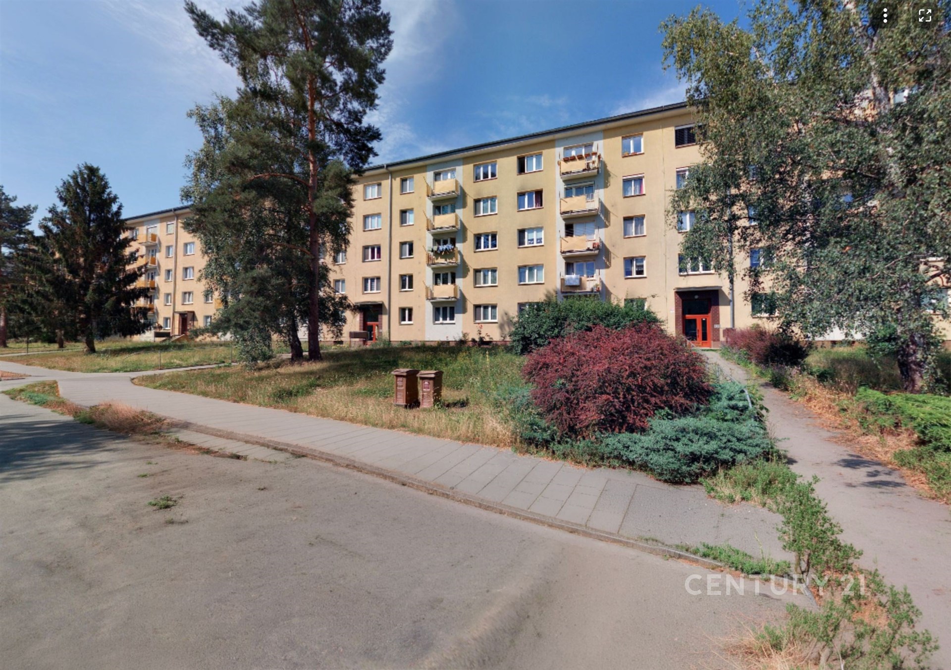 Čtyřpodlažní bytový dům s balkony, obklopený zelení a stromy, na klidném místě. Fasáda je v dobrém stavu, nabízí pohodlné bydlení v příjemné lokalitě.