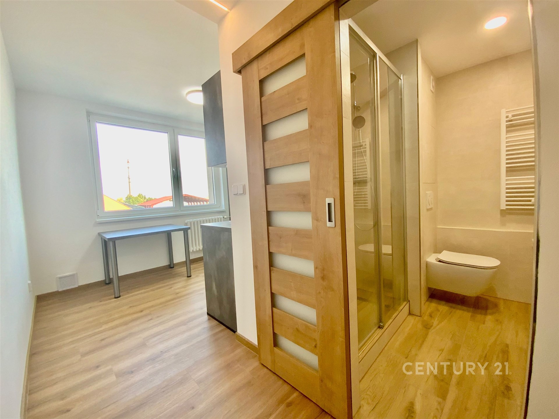 Rekonstruovaná koupelna s dřevěnými posuvnými dveřmi a moderní sprchou, vedle světlého pokoje s parketovou podlahou a velkými okny.