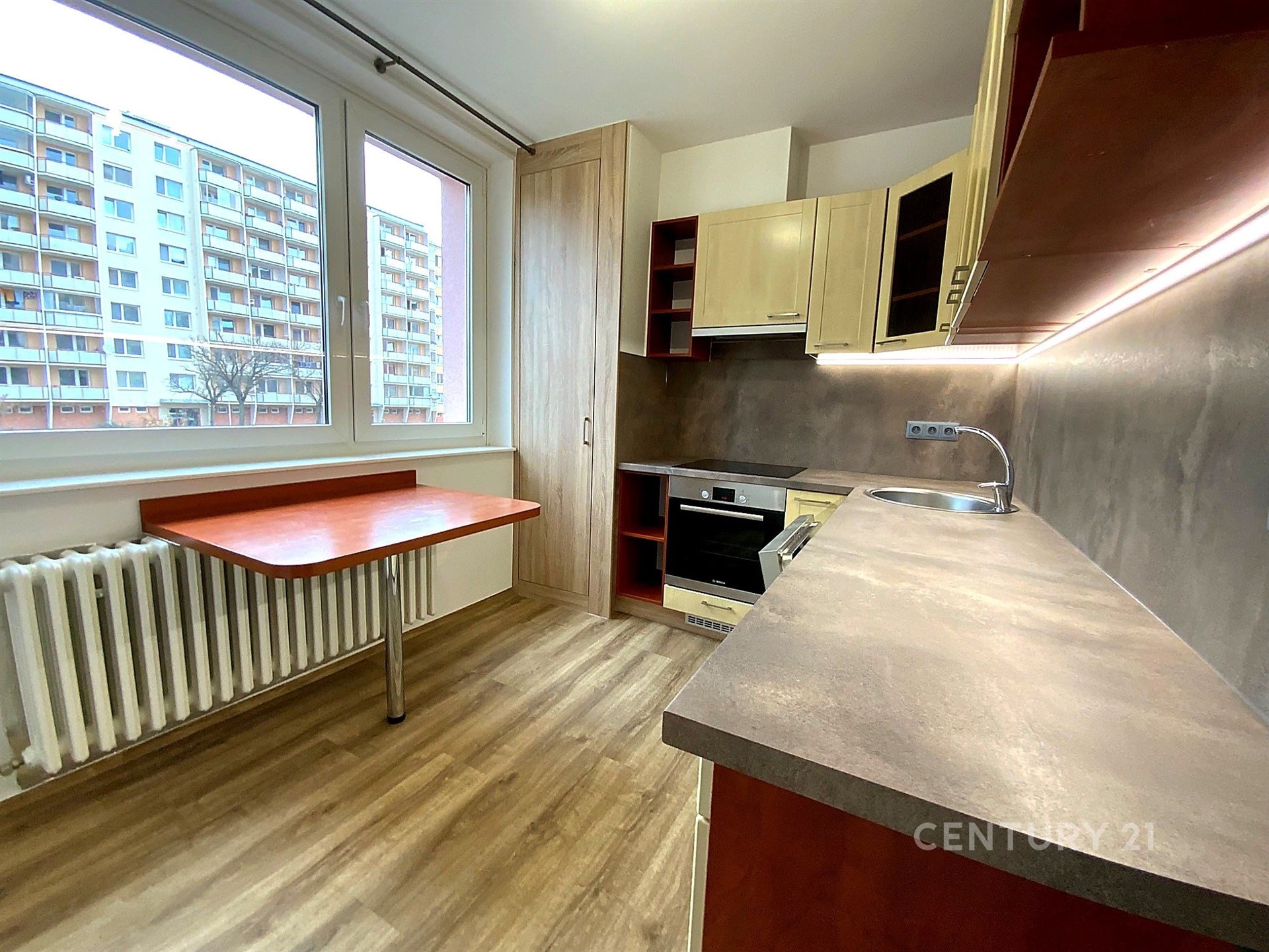 Moderní kuchyň s parketovou podlahou, vestavěným sporákem a velkými okny, poskytuje světlý prostor v rekonstruovaném bytě.