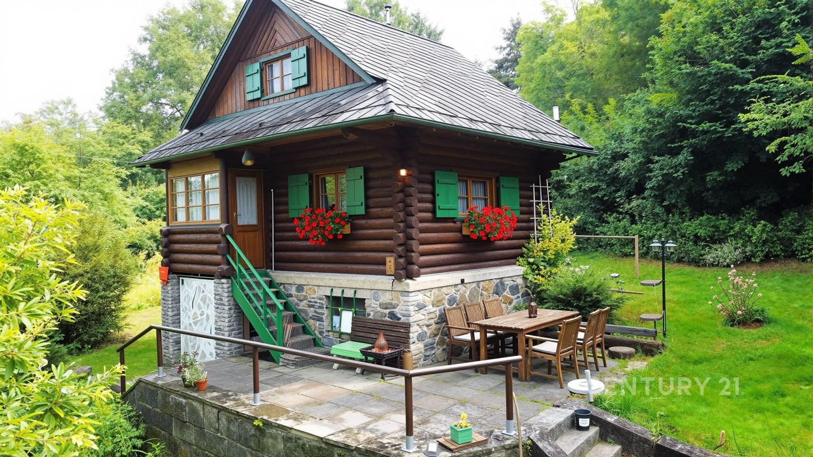 Dřevěná chalupa s kamenným základem, terasa s venkovním posezením, obklopená zelení, rustikální styl, klidná lokalita v přírodě.