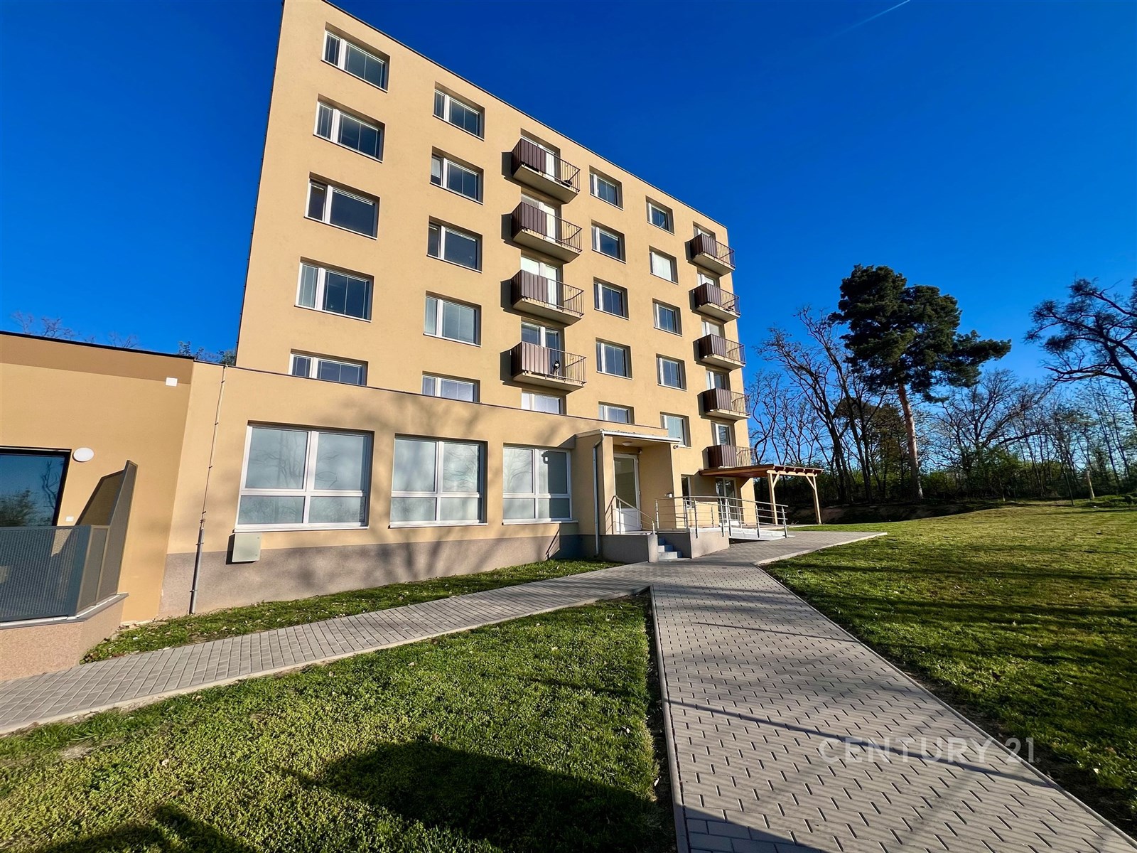 Moderní bytový dům s velkými okny, rekonstruovanou fasádou a prostornými balkony ve světlém odstínu, obklopený zelení a zpevněným chodníkem.