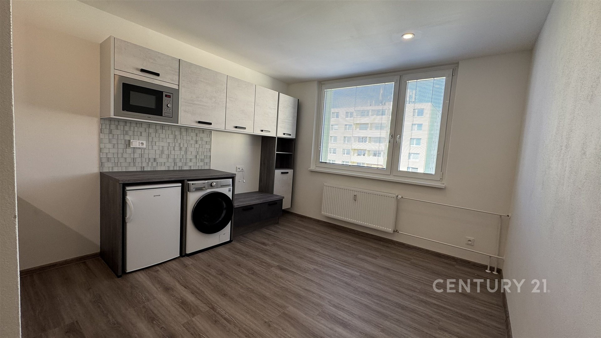 Kompaktní moderní kuchyň s dlažbou a vestavěnými spotřebiči, velké okno zajišťuje světlo a výhled, světlé stěny dodávají prostoru vzdušnost.