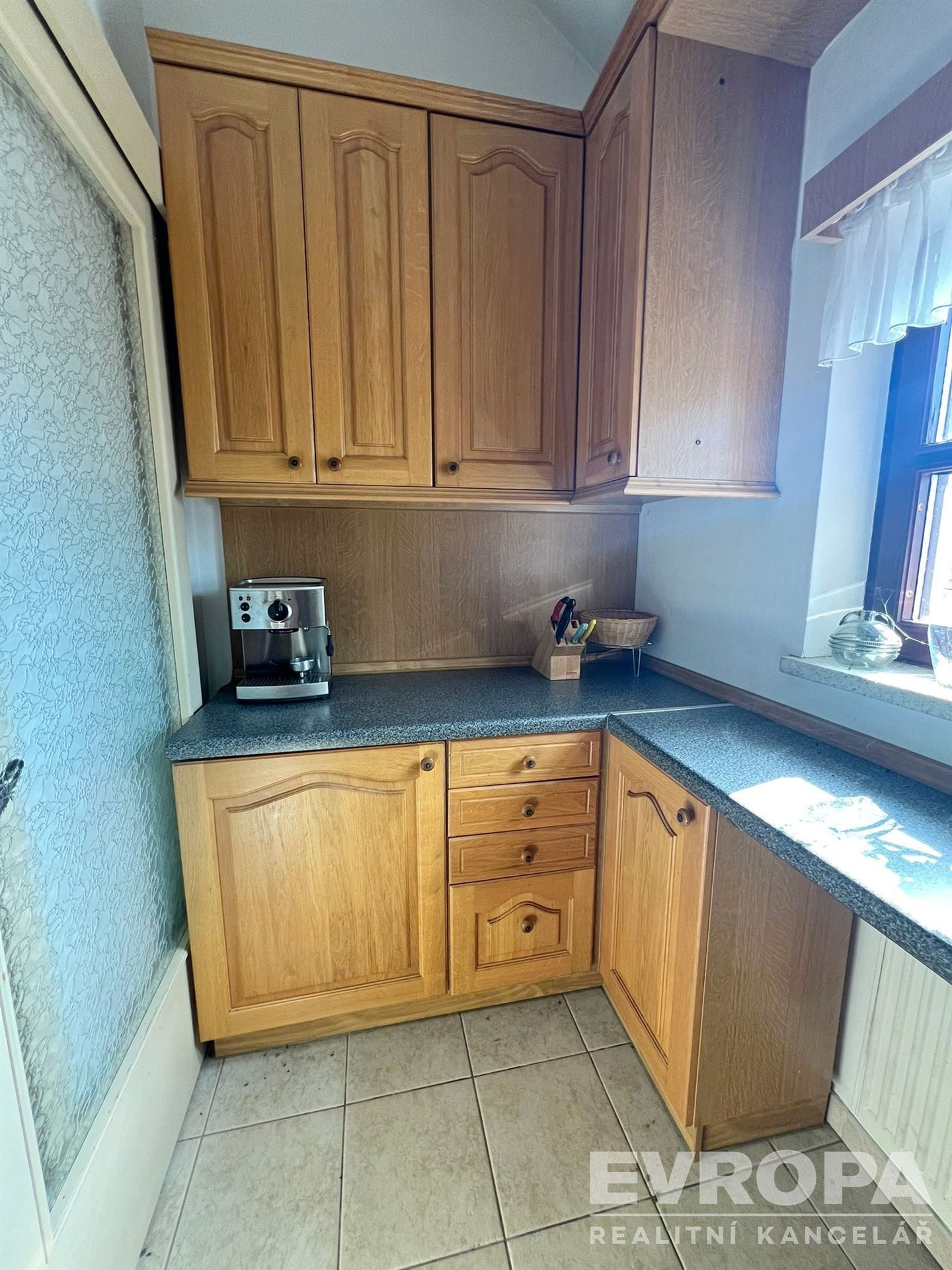 Kuchyň s dřevěnými skříňkami, moderními spotřebiči a dlažbou. Velká okna poskytují světlost a pohled na okolní krajinu.