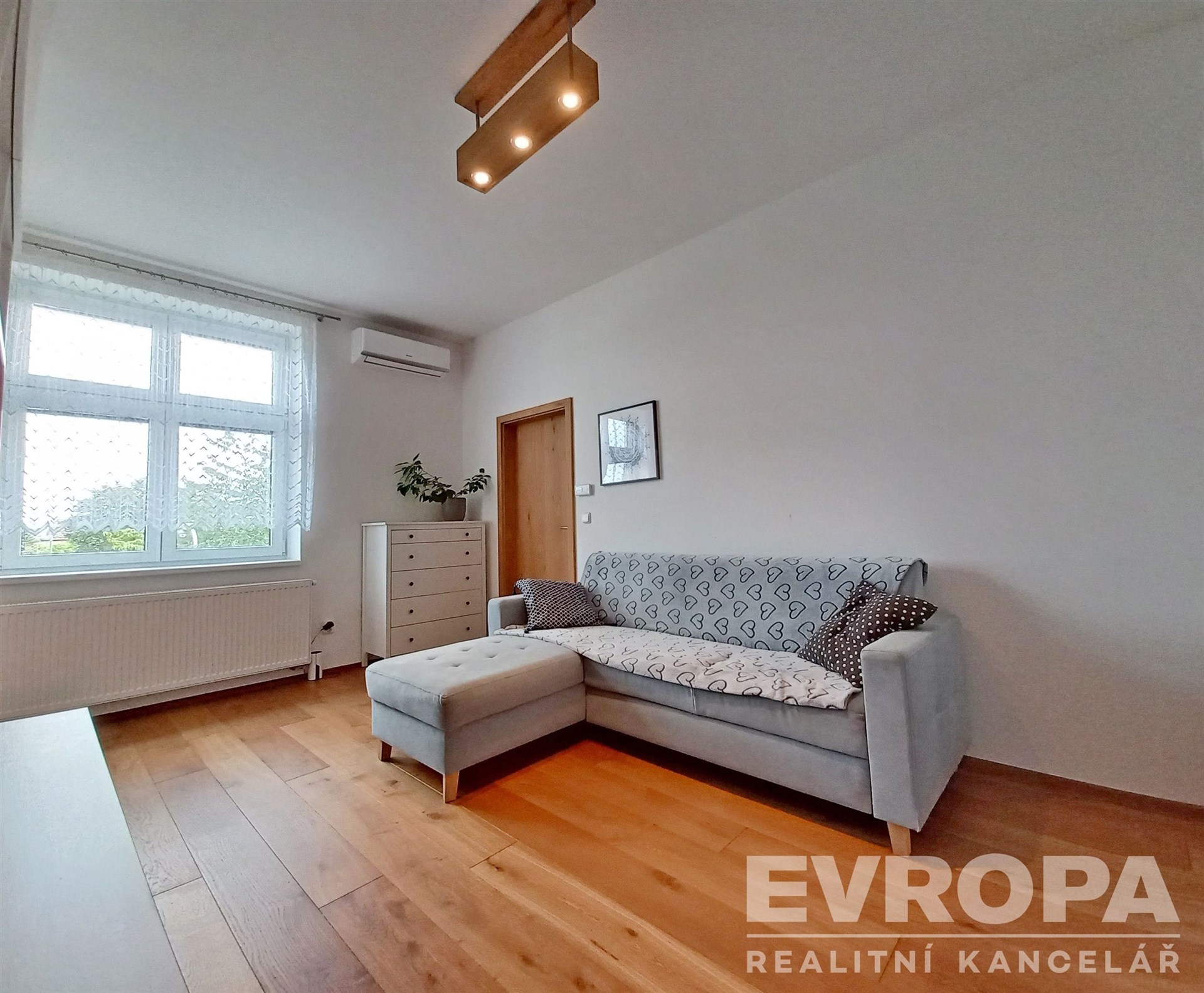 Světlý obývací pokoj s parketovou podlahou, velkými okny a moderním osvětlením, nabízí pohodlný prostor s klimatizací a minimalistickým designem.