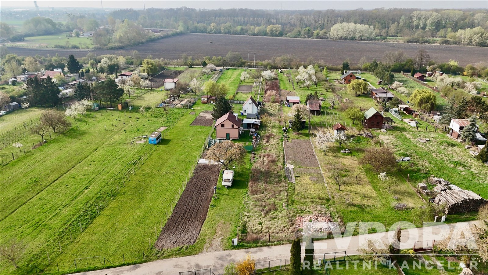Letecký pohled na rozlehlou zahradu s travnatými plochami a několika malými stavbami, obklopenou zelení a blízkým lesem.