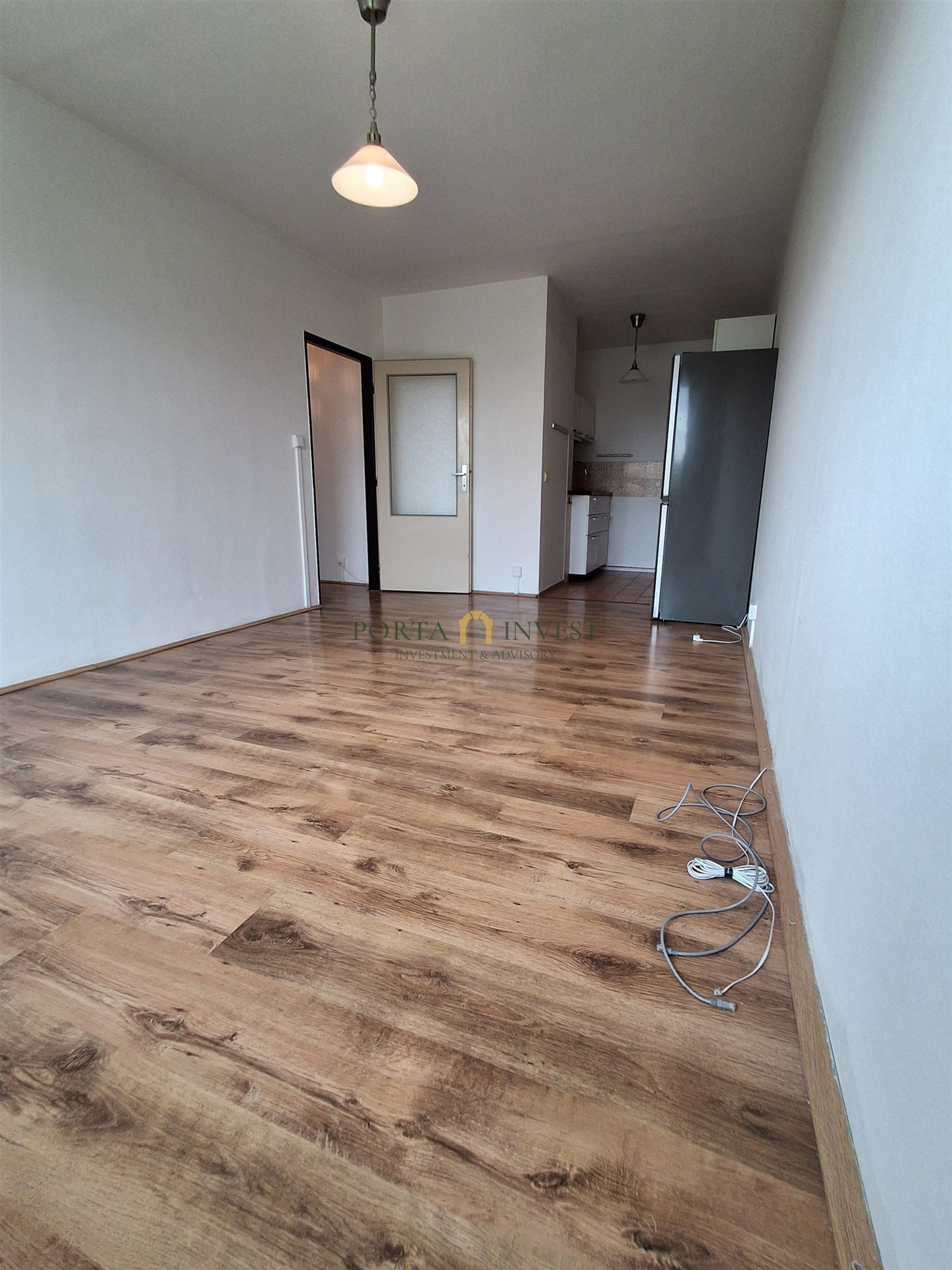 Prázdný obývací pokoj s parketovou podlahou, světlý interiér s moderním stylem, v pozadí kuchyňská linka.