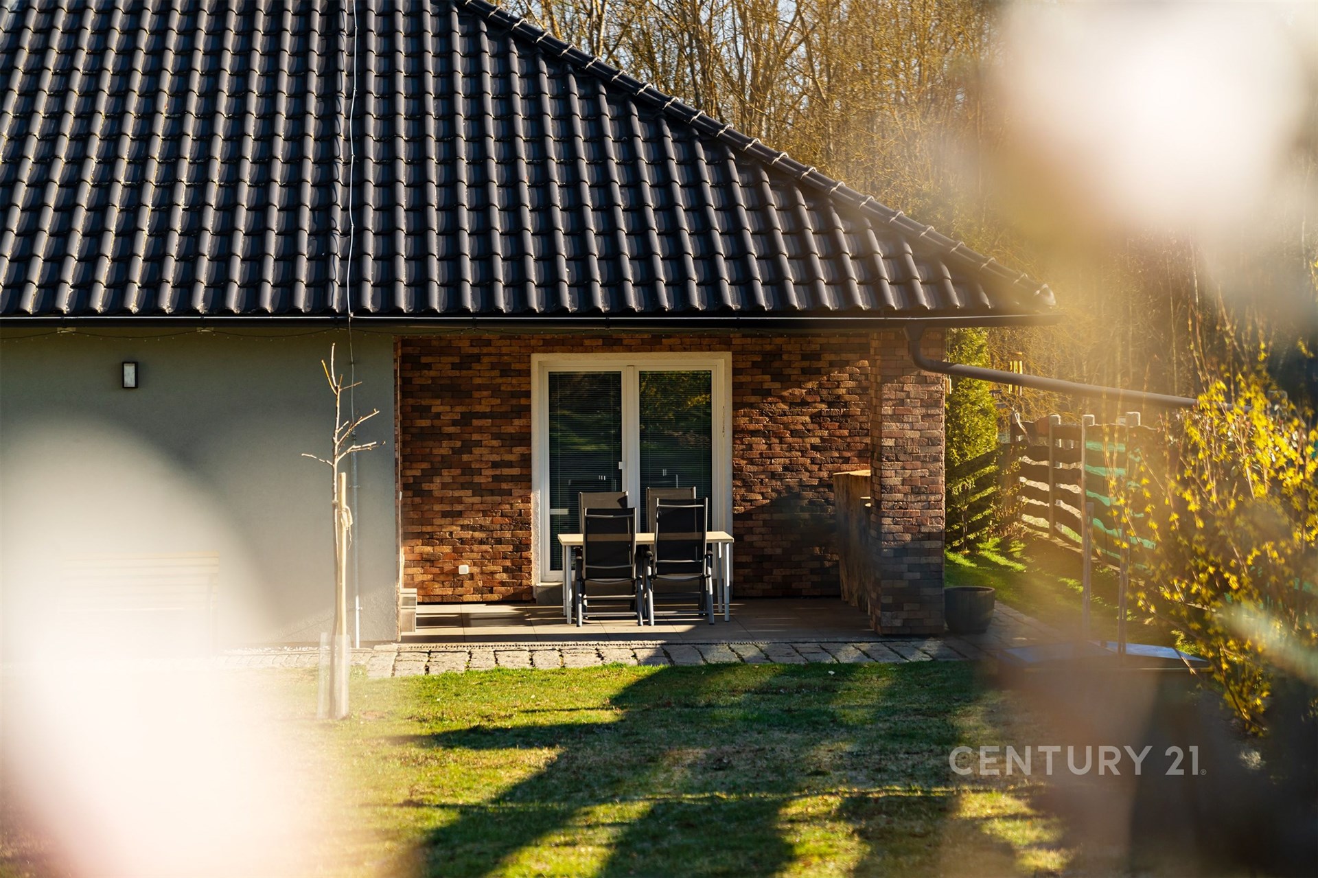 Exteriér domu s terasou, cihlovým obkladem a velkými okny, moderní styl. Udržovaná zahrada dodává příjemnou atmosféru.