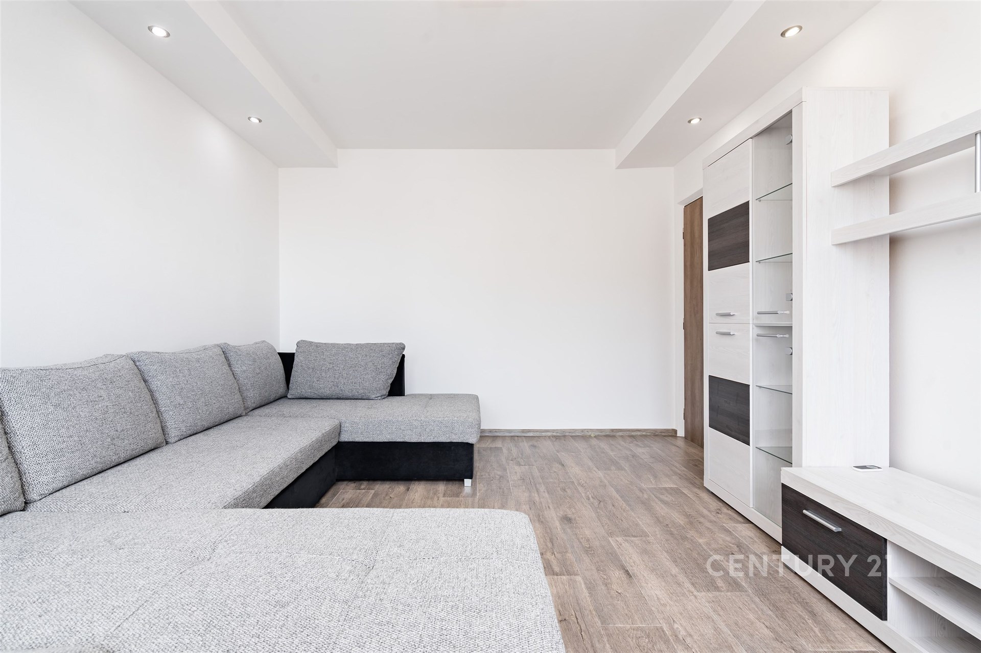 Světlý obývací pokoj s moderní sedací soupravou, laminátovou podlahou a vestavěnými policemi, minimalistický design a elegantní osvětlení.