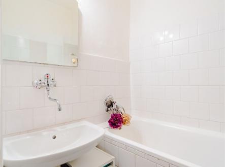 Koupelna s bílými kachličkami a vanou, moderní osvětlení, vestavěná umyvadlová skříňka, světlý prostor, příjemný minimalistický design.