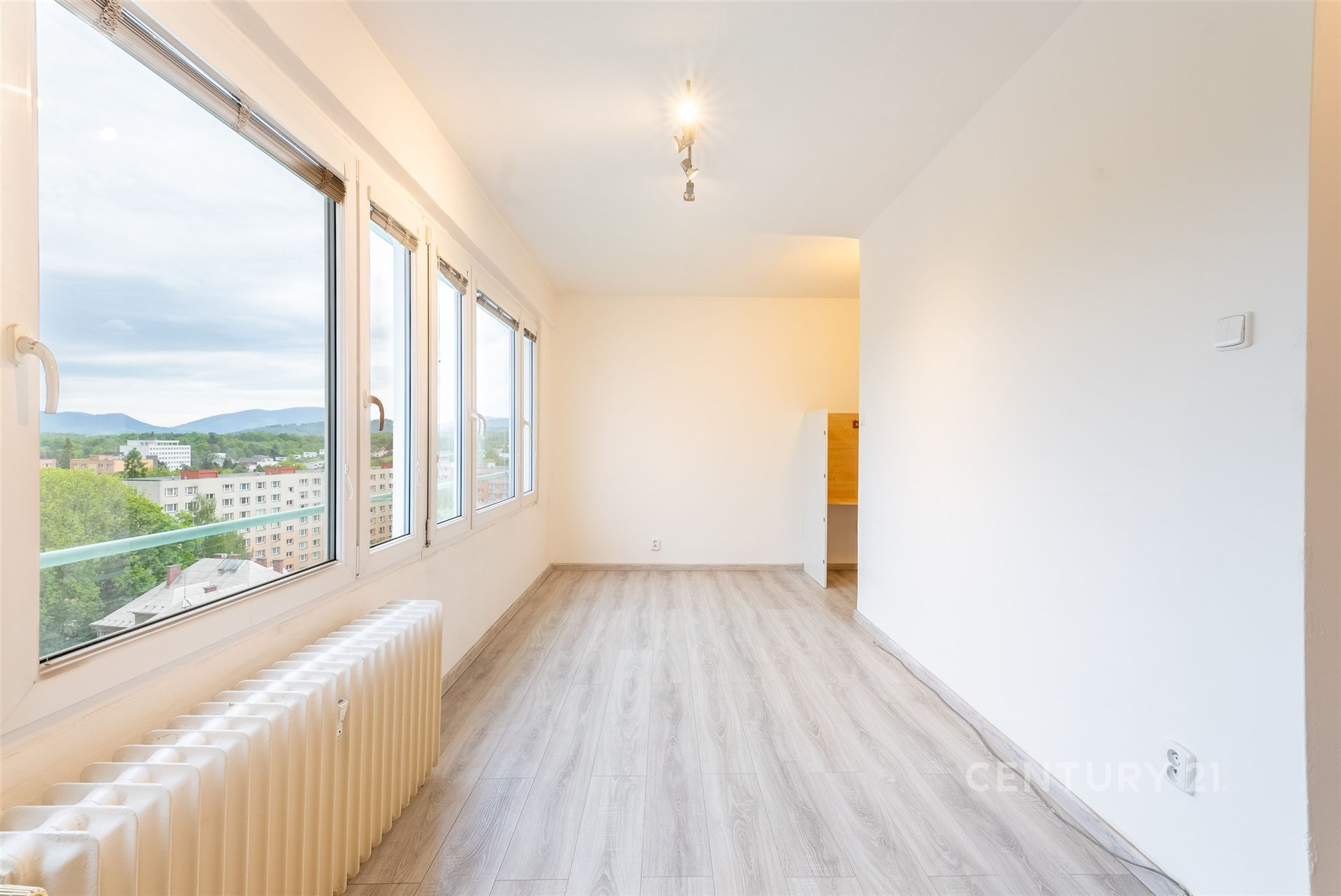 Světlý obývací pokoj s velkými okny, moderní parketová podlaha, výhled na město, minimalistické bílé stěny, prostorný vzhled.