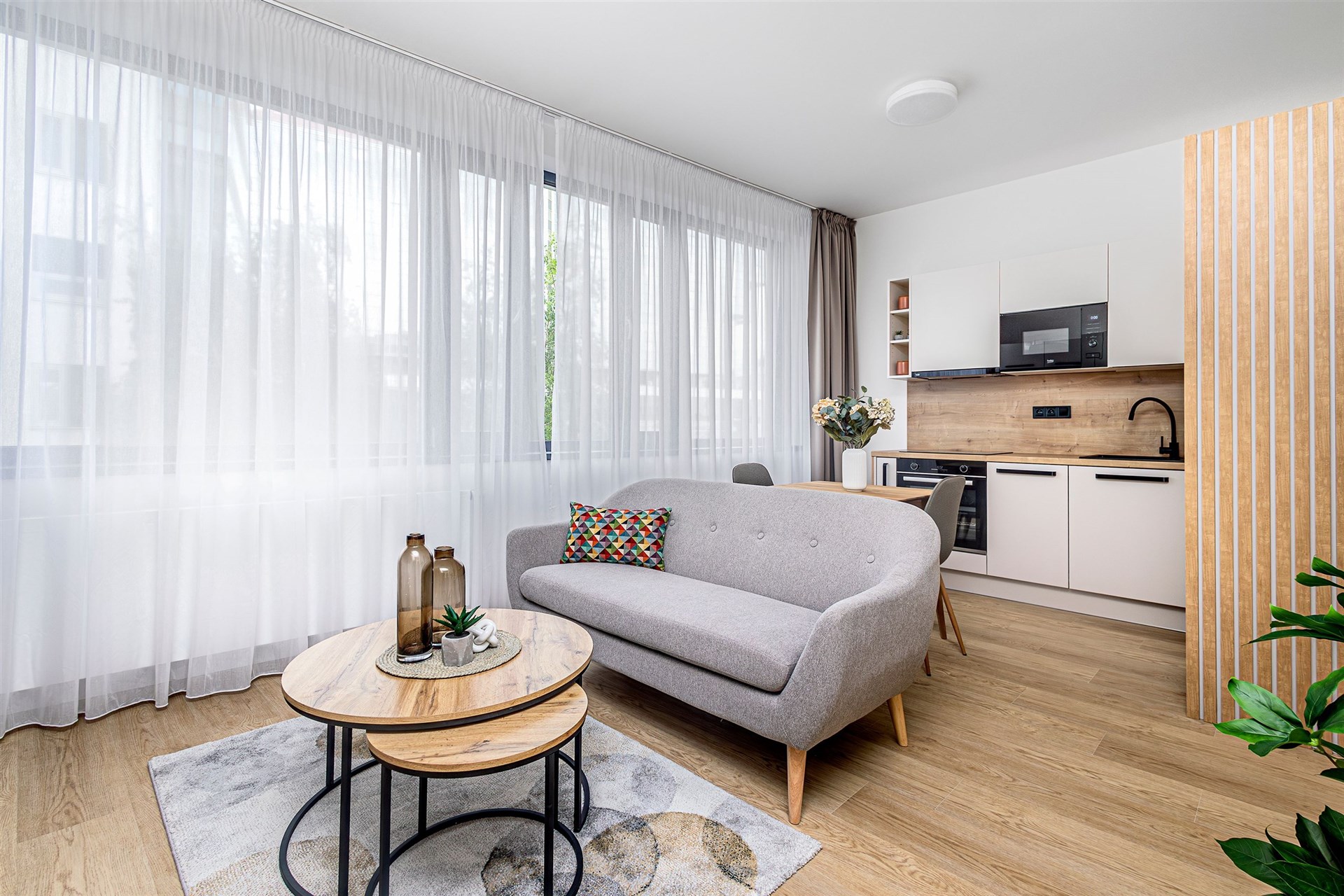 Světlý obývací pokoj s moderní kuchyní, parketová podlaha, velká okna s bílými záclonami, elegantní nábytek, světlý a vzdušný interiér.