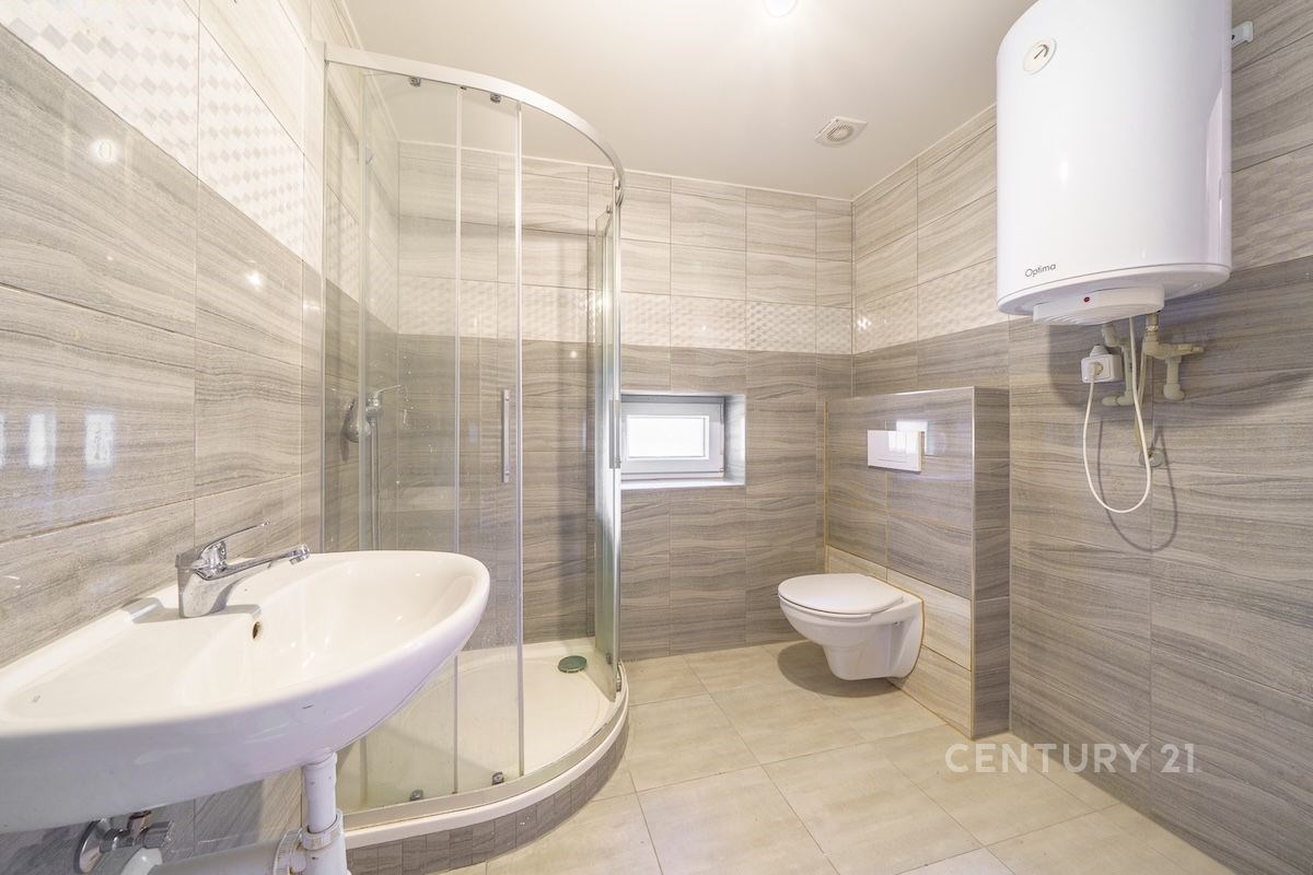 Prostorná koupelna s moderními obklady, sprchovým koutem a závěsnou toaletou, světlá dlažba, bojler na stěně.