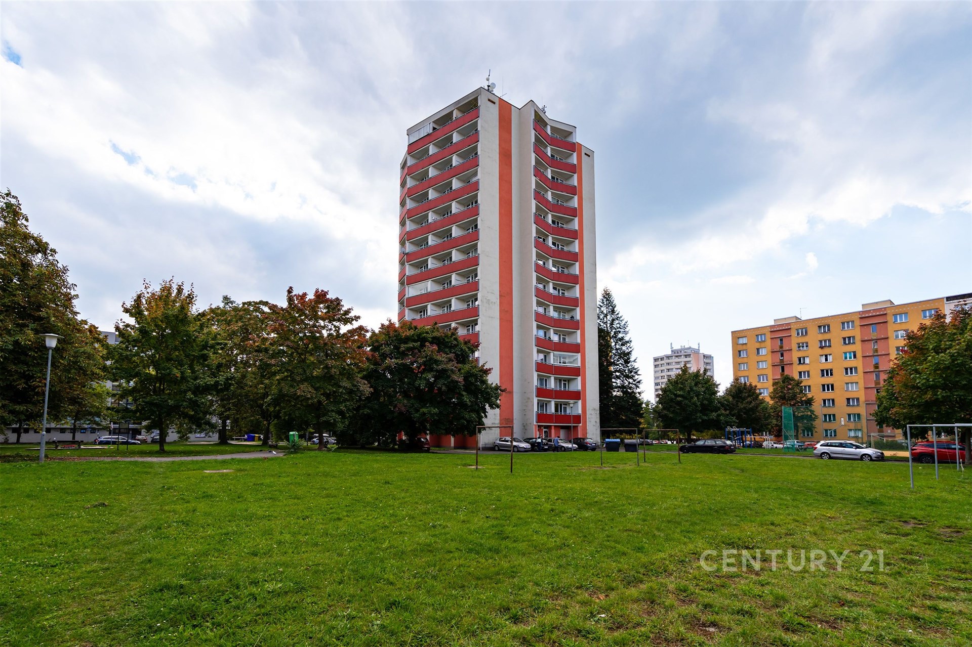 Vysoký bytový dům s balkony, obklopený zeleným parkem a stromy, poblíž parkování. Budova je moderní a nachází se v klidné lokalitě.