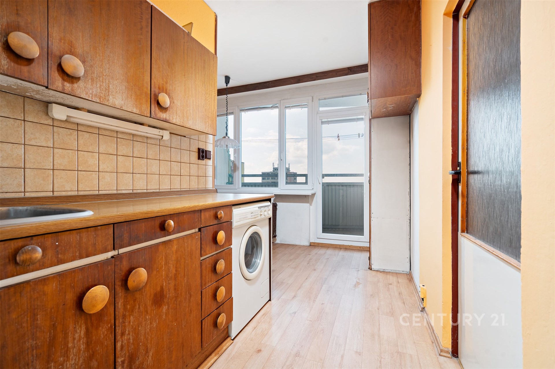 Kuchyň s dřevěnými skříněmi, prostor pro pračku a velkým oknem. Dřevěné prvky a světlá laminátová podlaha dodávají místnosti útulnost.