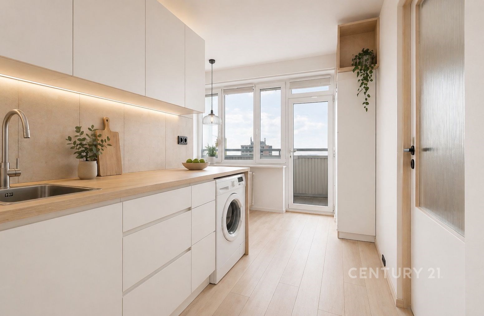 Světlá moderní kuchyň s dřevěnou pracovní deskou, bílými skříňkami a parketovou podlahou, velká okna s balkonovými dveřmi poskytují dostatek světla.