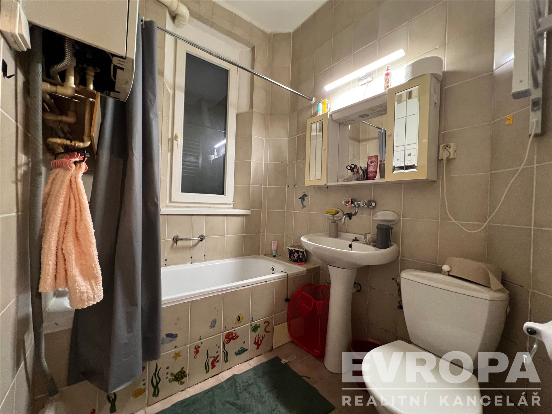 Koupelna s dlažbou, vanou a závěsnou skříňkou, obsahuje velké okno a kombinaci světlých obkladů, působí udržovaně a funkčně.