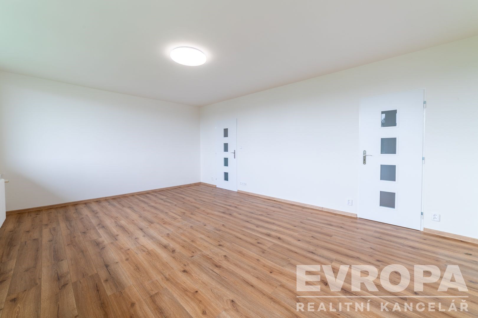 Světlý obývací pokoj s parketovou podlahou, moderní dveře a minimalistický styl, prostorný interiér vhodný k osobnímu uspůsobení.