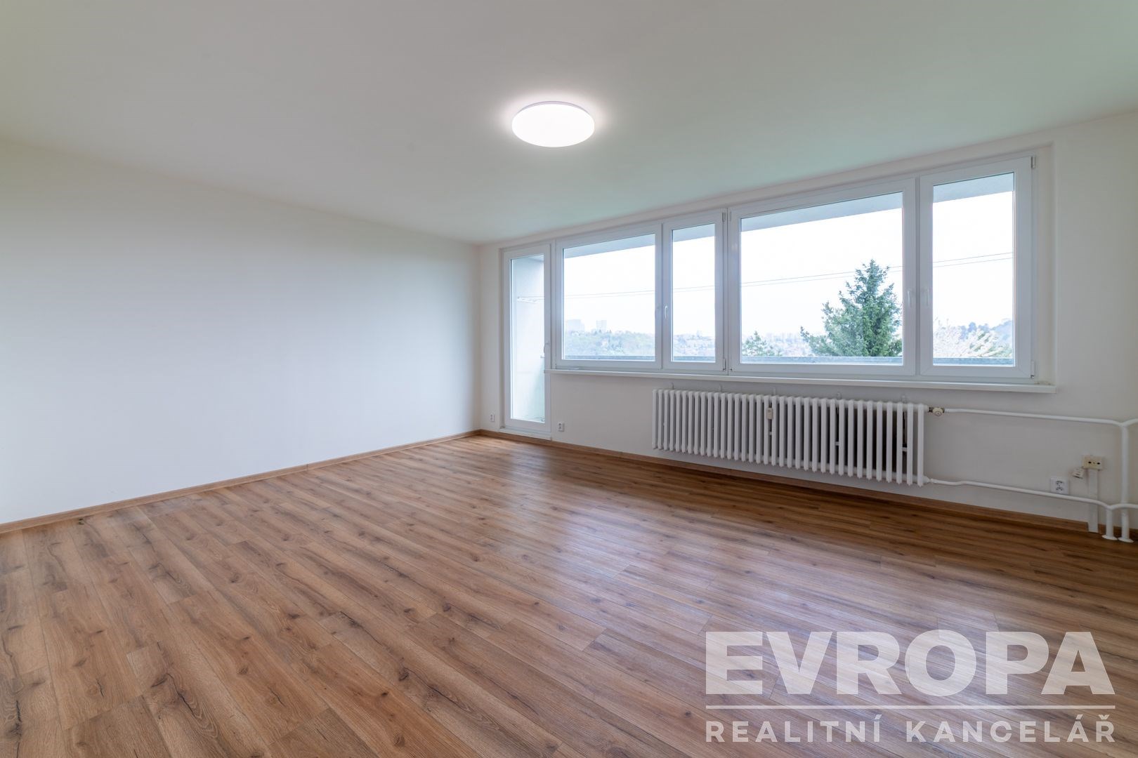 Světlý obývací pokoj s parketovou podlahou a velkými okny nabízí moderní prostor s výhledem, ideální pro pohodlné bydlení.