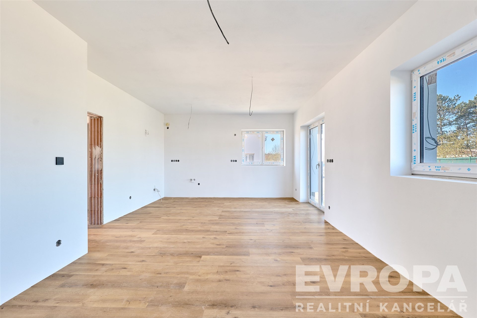 Světlý obývací pokoj s parketovou podlahou, velkými okny a moderním stylem, ideální pro rodinné bydlení.