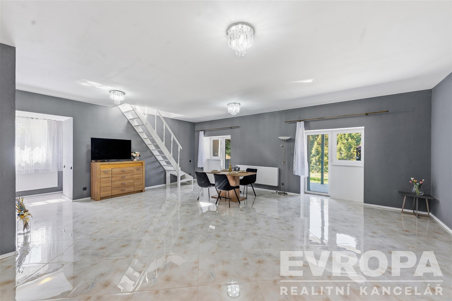 Prostorný obývací pokoj s velkými okny, moderní dlažbou a schodištěm; interiér nabízí světlý, elegantní prostor ideální pro rodinné bydlení.