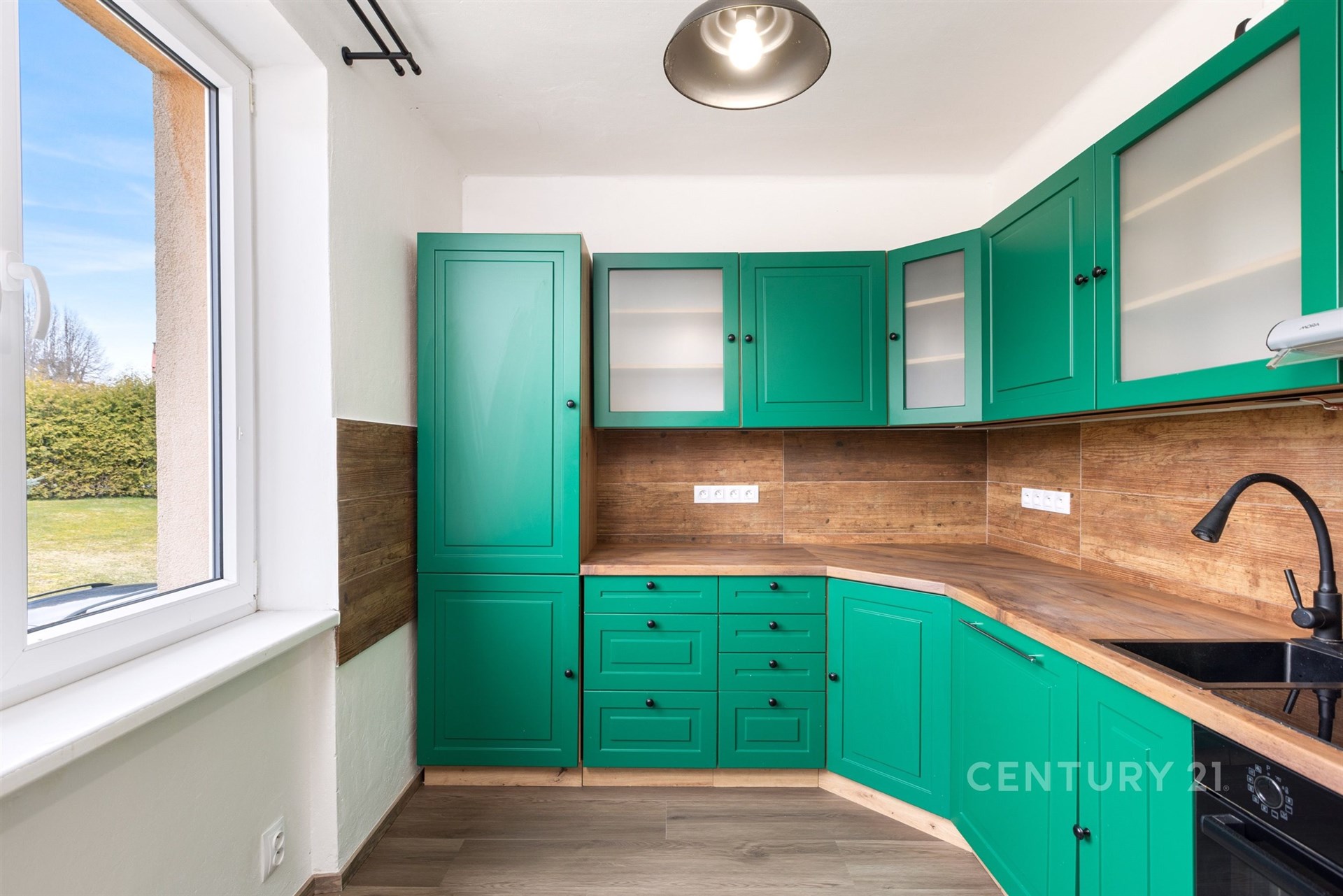 Kuchyň s dřevěnými prvky, moderními zelenými skříněmi a velkým oknem, dřevěný vzhled pracovní desky, světlý prostor.