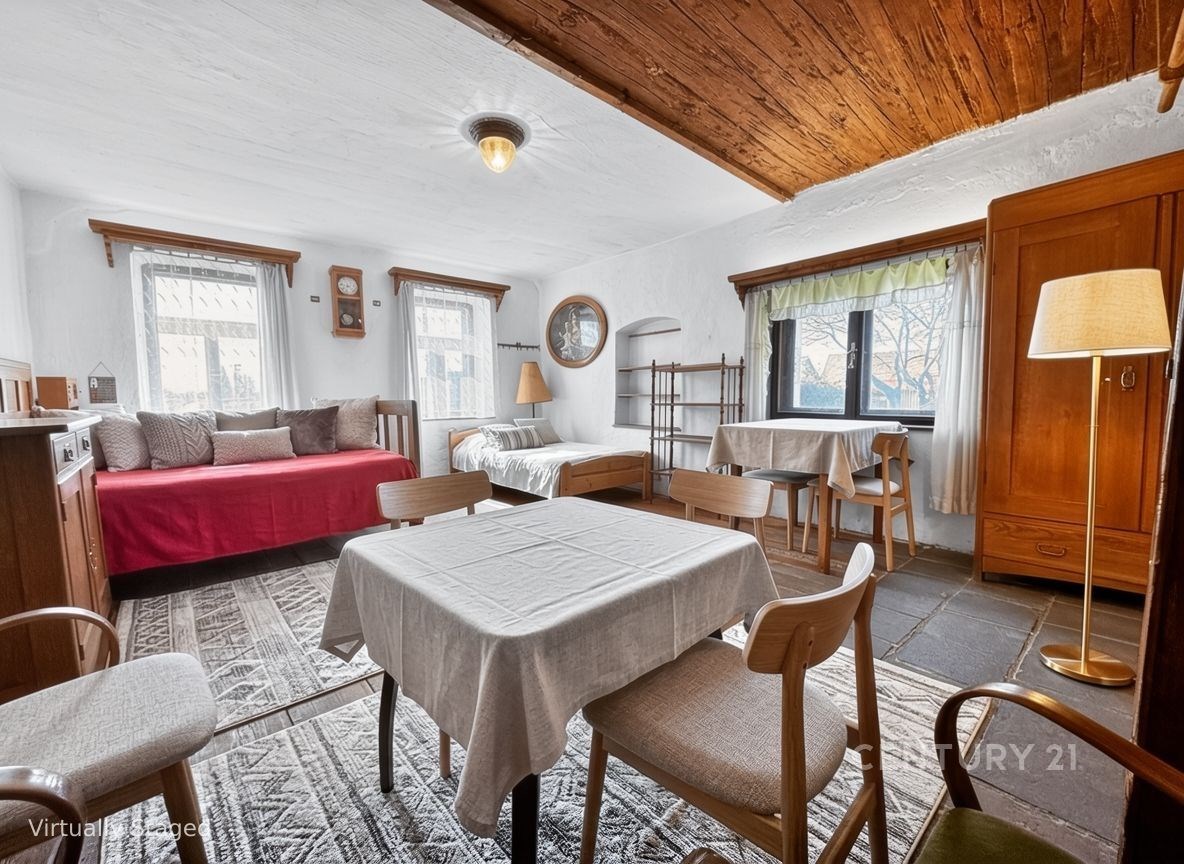 Světlý obývací pokoj s kamennou dlažbou, dřevěným stropem a velkými okny, obsahuje dřevěný nábytek a jídelní kout, působí útulně.