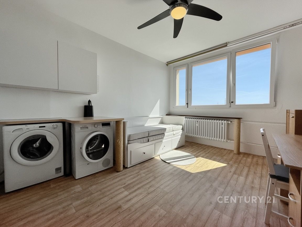 Světlý obývací pokoj s dřevěnou podlahou, velká okna, moderní interiér, v rohu místo pro praní s pračkou a sušičkou.