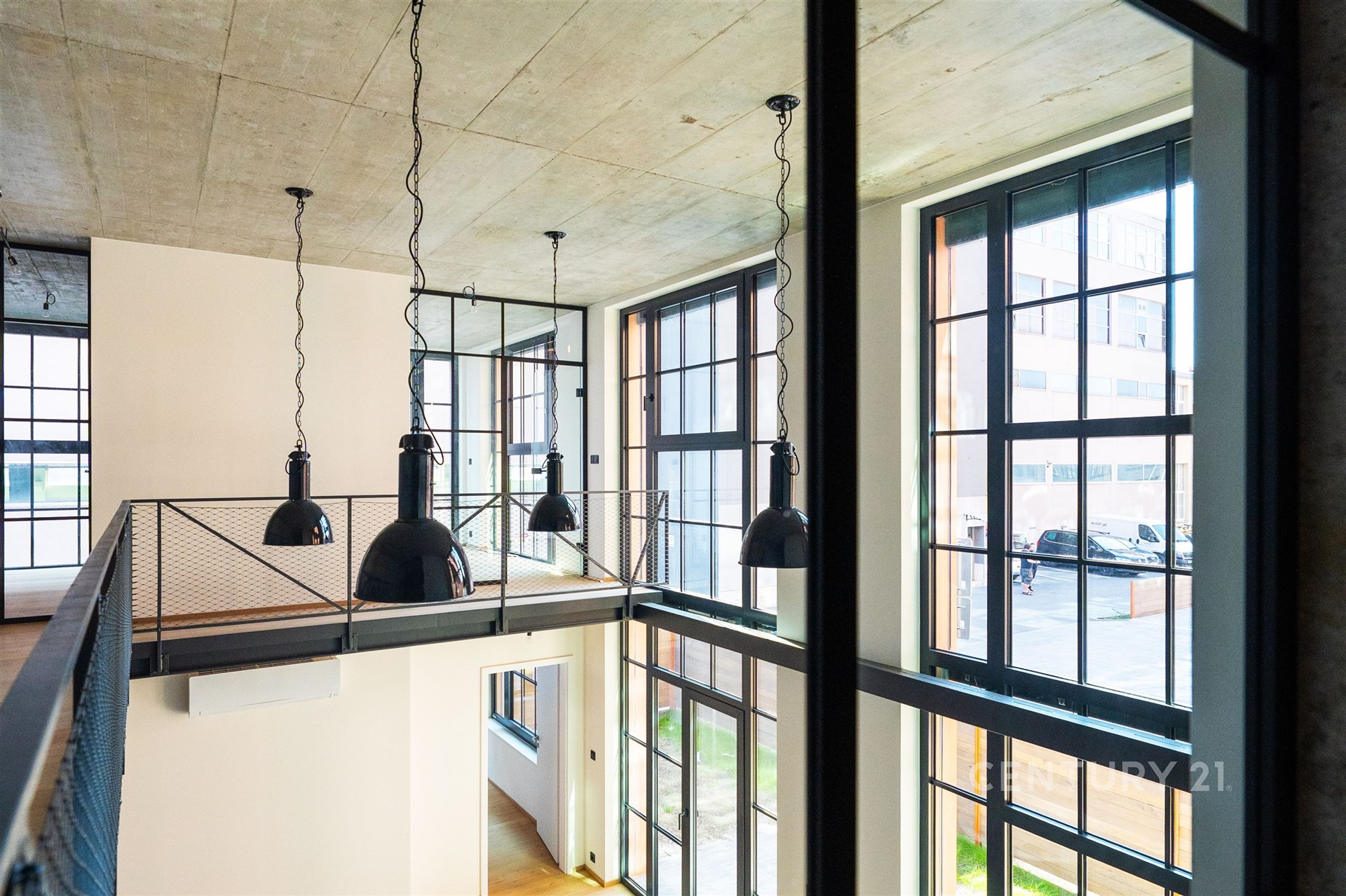 Moderní interiér s vysokým stropem, velká okna a parketová podlaha, industriální styl s kovovými prvky, ideální pro loftové bydlení.