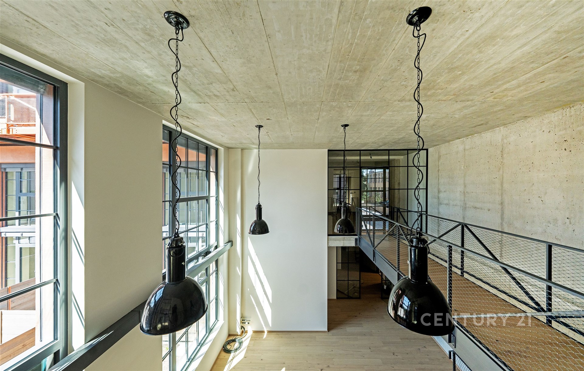 Industriální obývací pokoj s betonovým stropem, velkými okny a dřevěnou podlahou, moderní loft styl a kovové zábradlí.