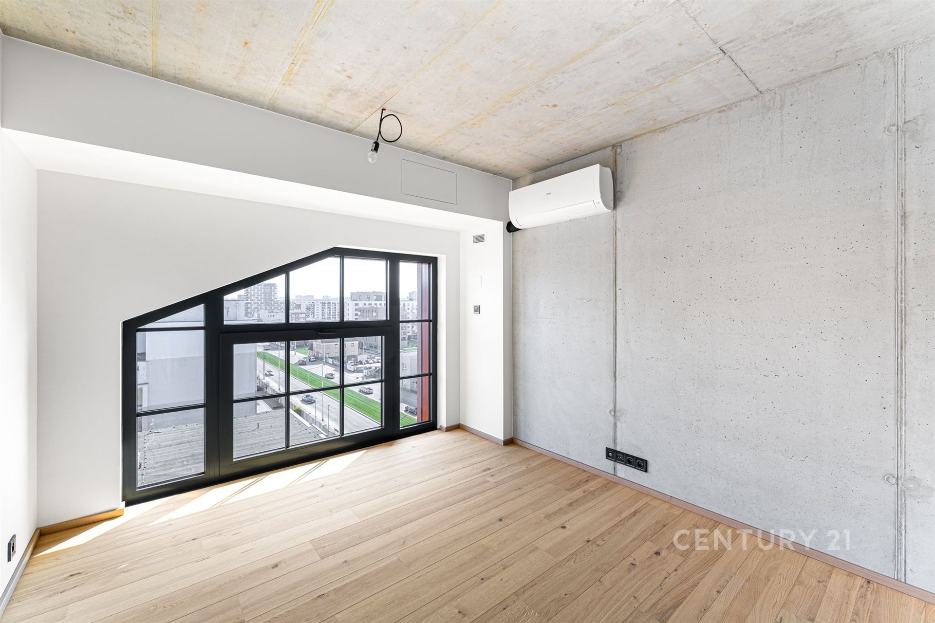 Světlý obývací pokoj s parketovou podlahou a velkými okny, moderní klimatizace a betonové stěny vytvářejí stylový interiér.