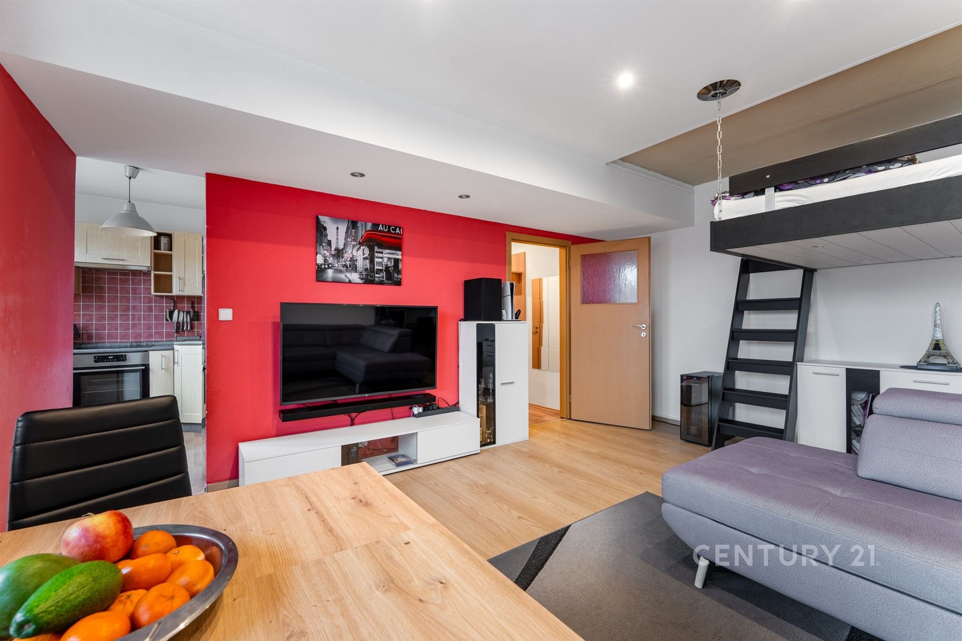 Obývací pokoj s moderním nábytkem, parketovou podlahou a zvýšeným spacím prostorem. Velké okno poskytuje dostatek světla, svěží dekorace.