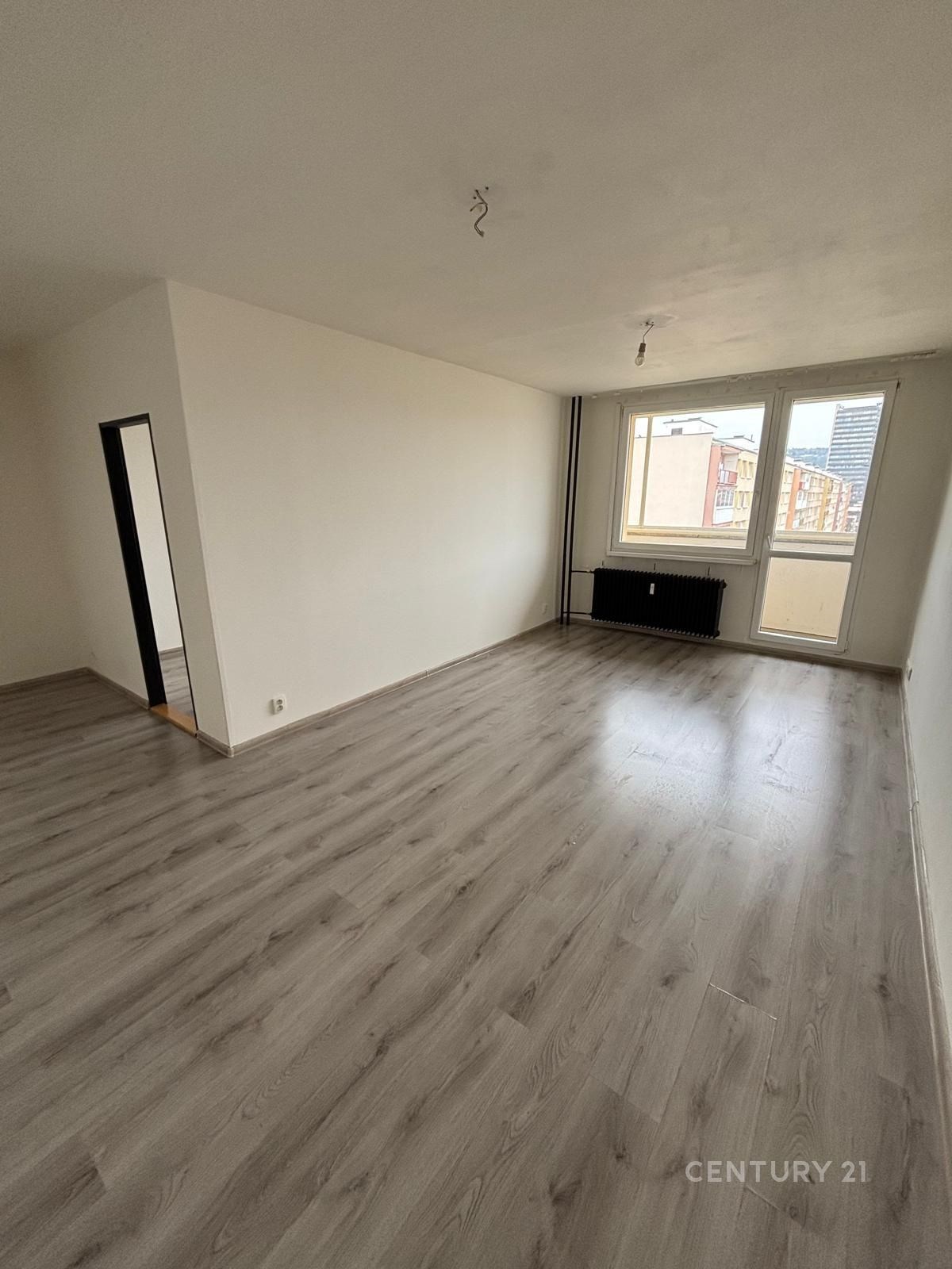 Světlý obývací pokoj s laminátovou podlahou, velká okna, světlý interiér, výhled na město, moderní styl, renovovaný prostor.