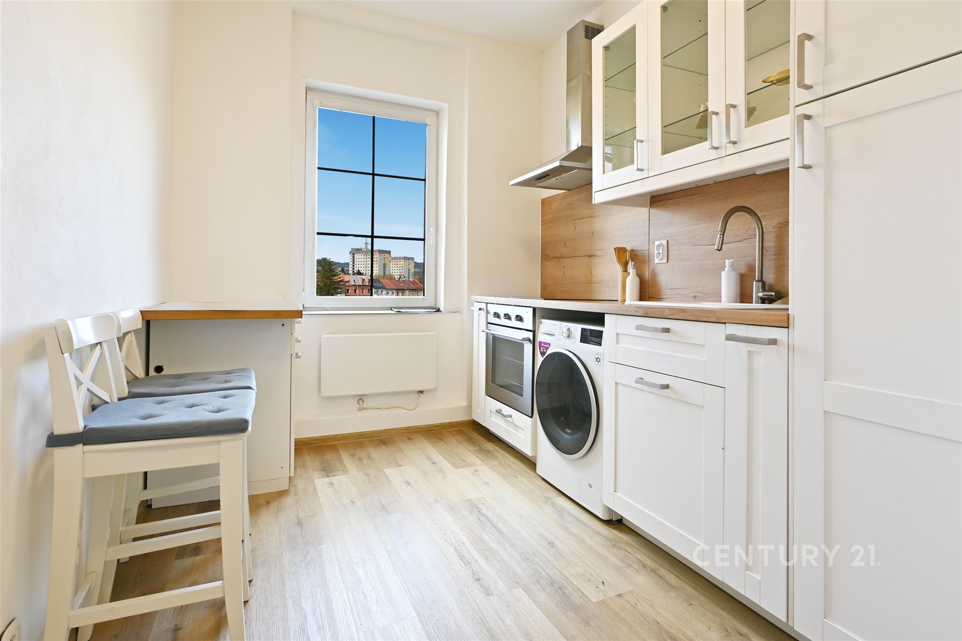 Kompaktní světlá kuchyň s moderními spotřebiči, parketová podlaha a bílé skříňky.