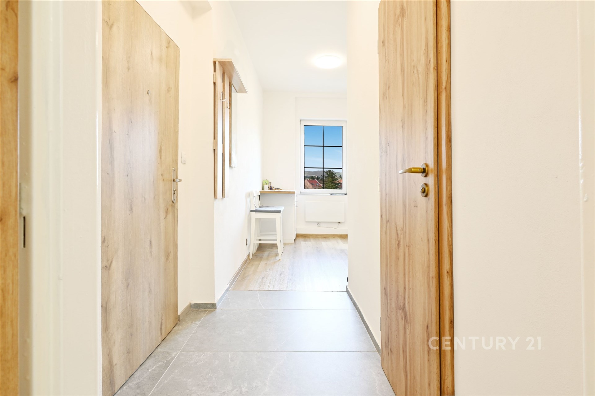 Světlá chodba s keramickou dlažbou, dřevěnými dveřmi a velkým oknem, moderní úprava interiéru, navazuje na obytný prostor.