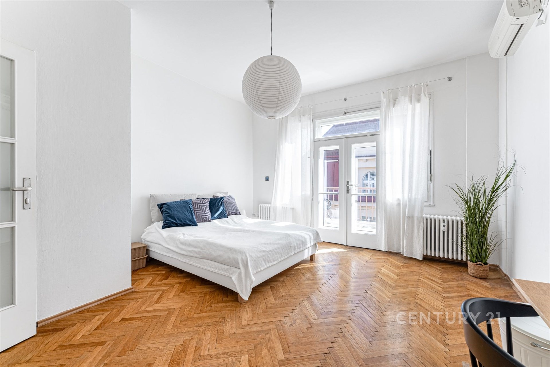 Prostorná ložnice s parketovou podlahou, velkými okny a moderní klimatizací, nabízí světlo a styl v rekonstruovaném bytě.