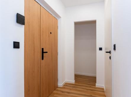 Úzká předsíň s moderní parketovou podlahou, dřevěnými dveřmi a bílými stěnami, ideální pro elegantní vstupní prostor.