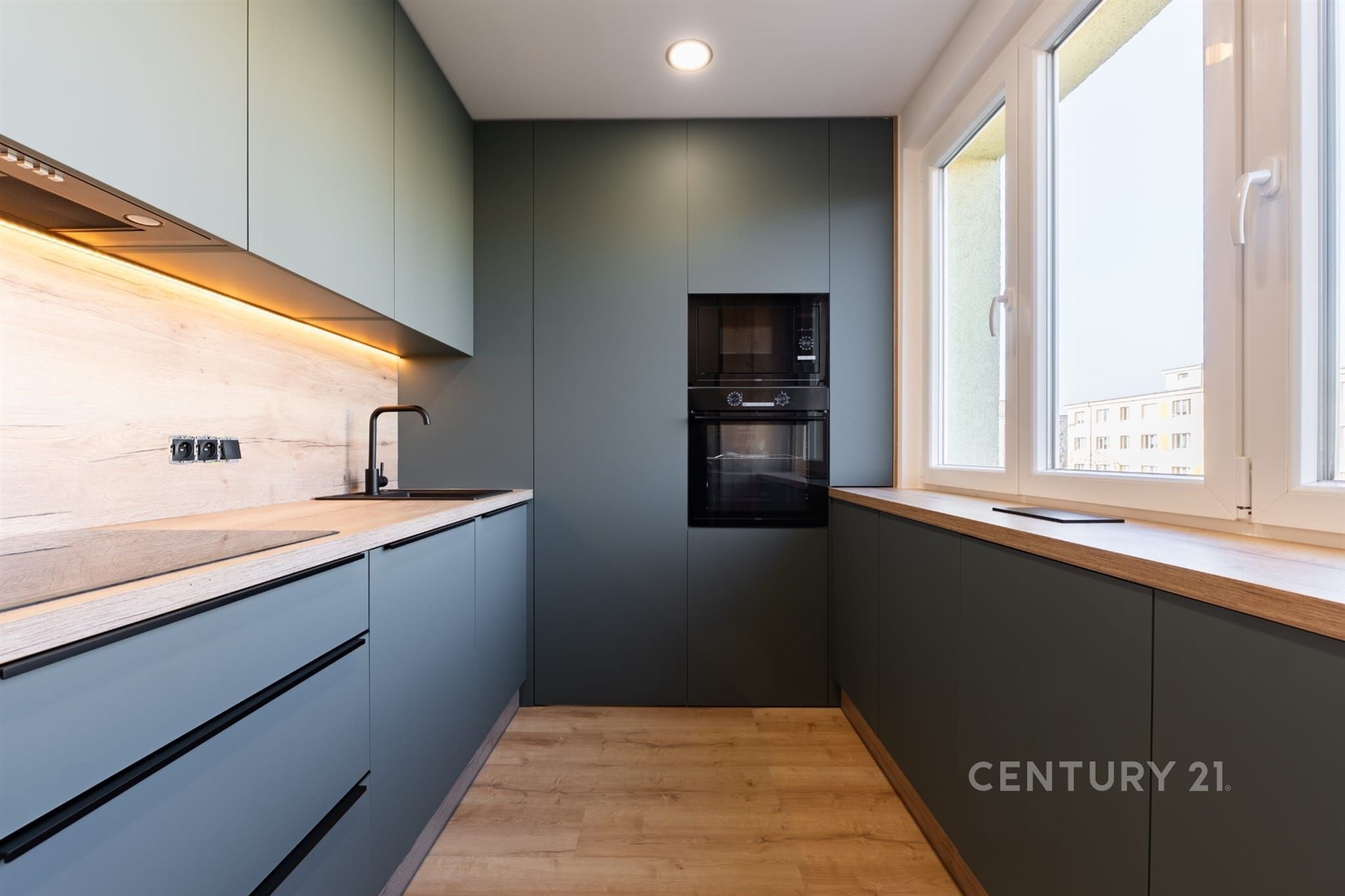 Moderní kuchyň s parketovou podlahou a vestavěnými spotřebiči, velká okna poskytují dostatek světla, elegantní a funkční design.