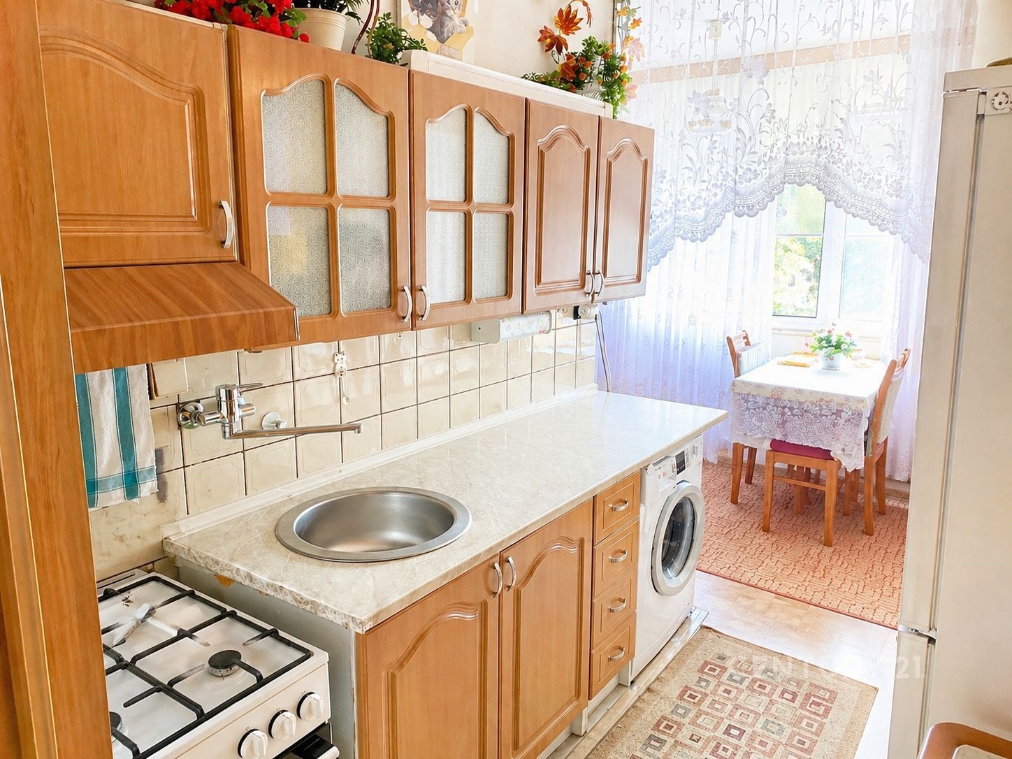 Kuchyň s dřevěnými skříněmi, bílou dlažbou a velkým oknem, které poskytuje dostatek světla, součástí je i malý jídelní kout.