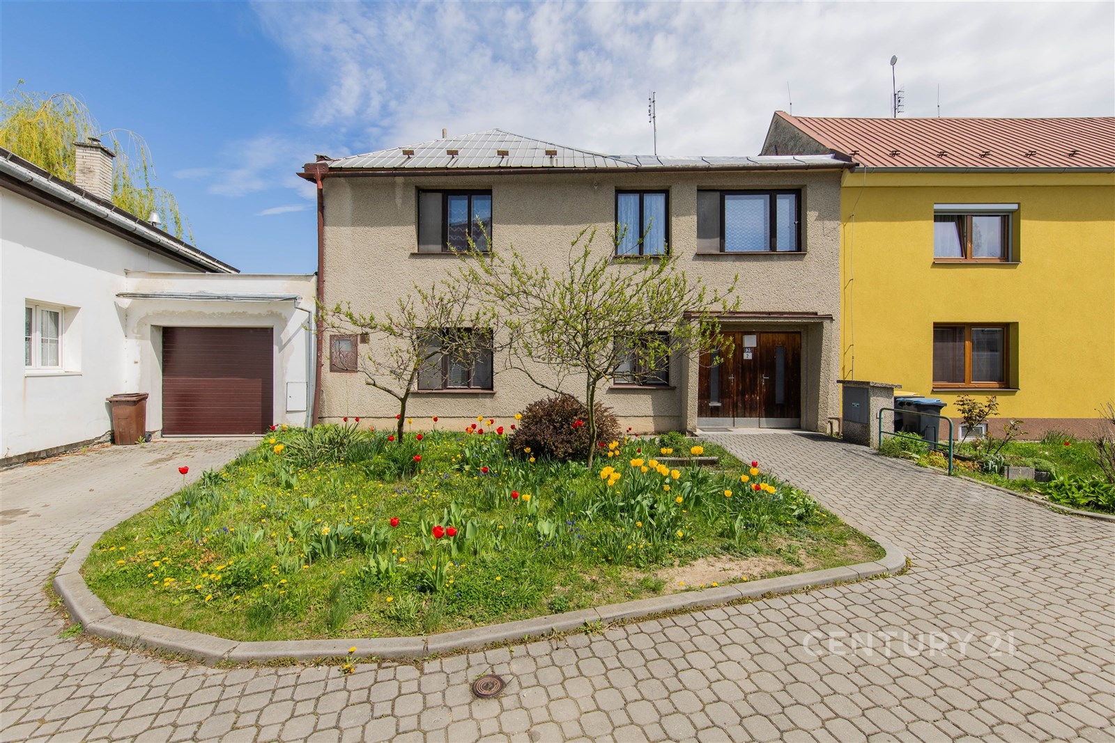 Rodinný dům s tradiční fasádou, upravenou zahradou a garáží. Přístupová cesta je z dlažby. Dům má moderní okna a udržovaný exteriér.