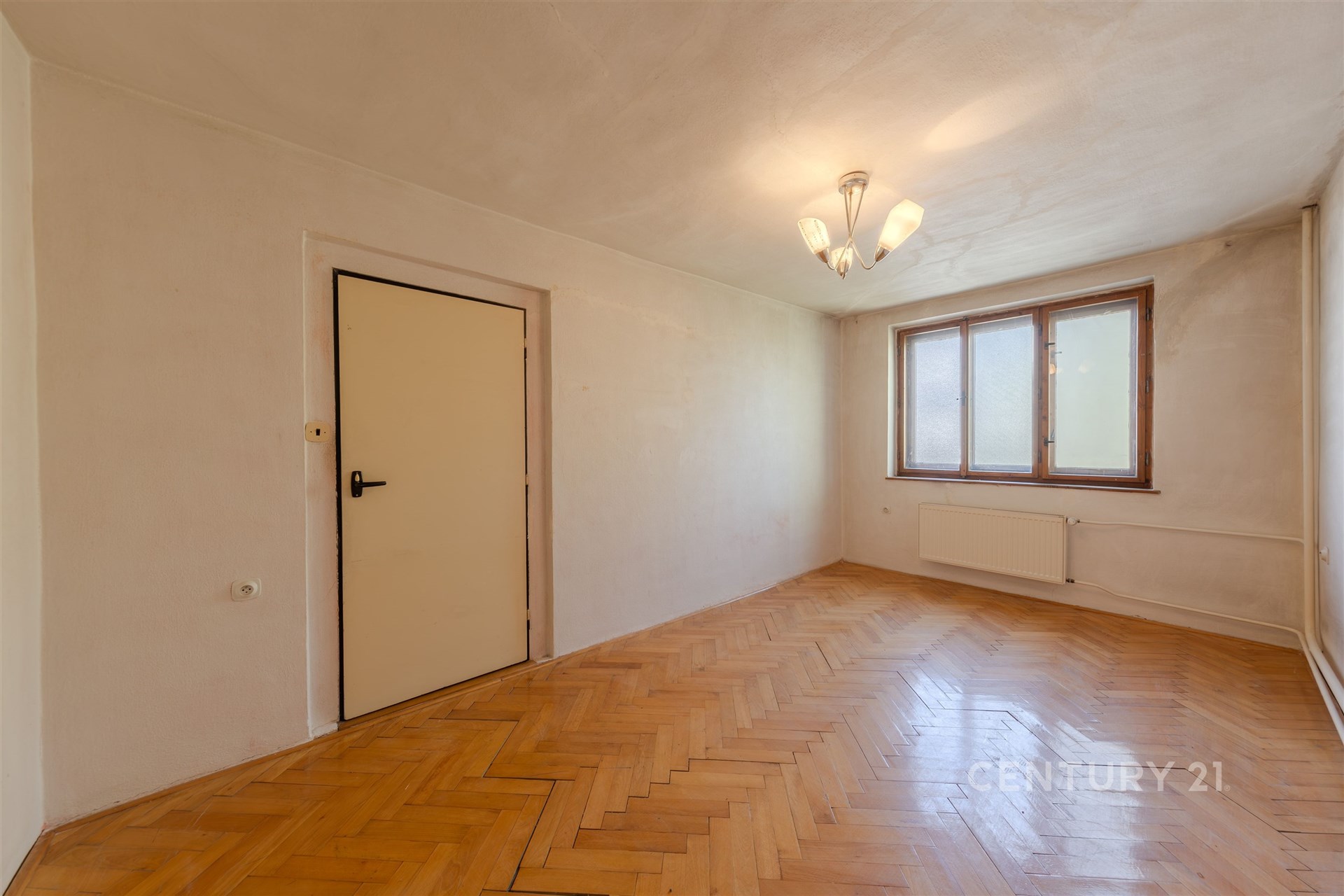 Světlý obývací pokoj s parketovou podlahou a velkými okny, moderní osvětlení a zdi připravené k renovaci.