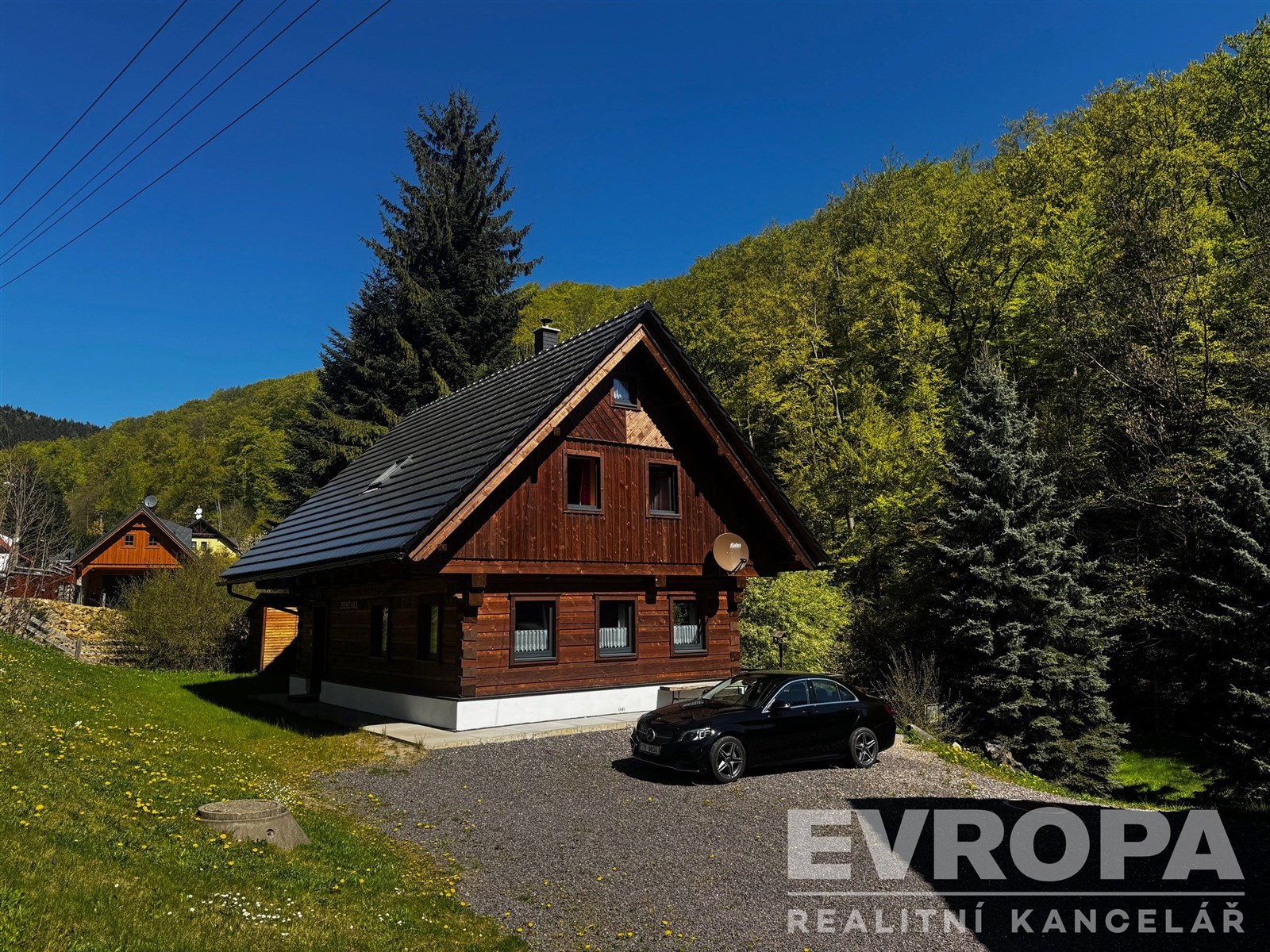 Dřevěná chalupa s moderní střechou, umístěná v klidném prostředí obklopeném zelení a vzrostlými stromy, ideální pro milovníky přírody.