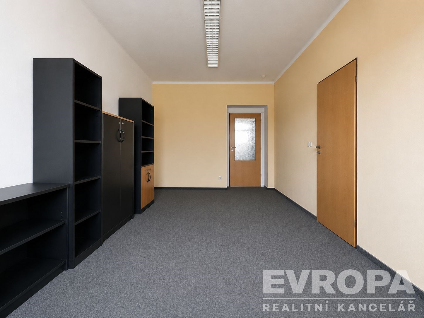 Prázdný kancelářský prostor s kobercem, světlými stěnami a dřevěnými dveřmi, vestavěné skříně, moderní osvětlení, vhodný pro komerční využití.