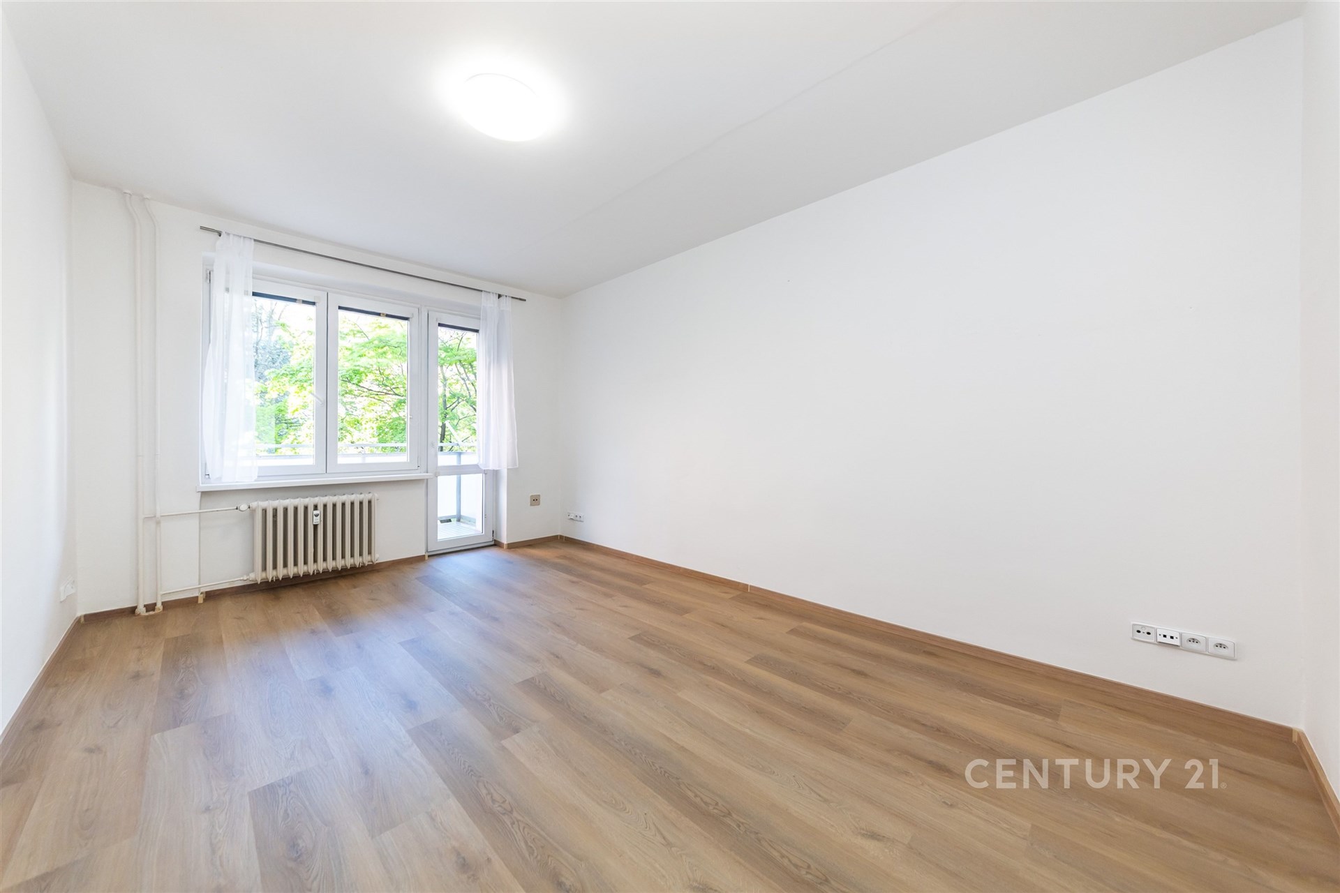 Světlý obývací pokoj s parketovou podlahou a velkými okny, moderní prostor s bílými stěnami vhodný k okamžitému nastěhování.