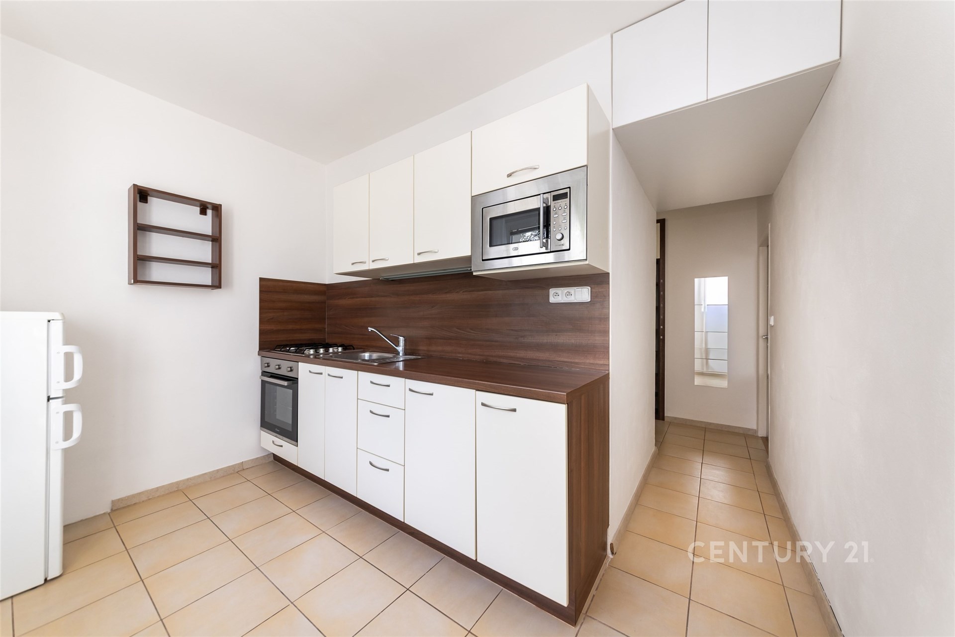 Kompaktní kuchyň s moderními bílými skříňkami, vestavěnou mikrovlnnou troubou a dlažbou. Světlý a prakticky zařízený prostor.