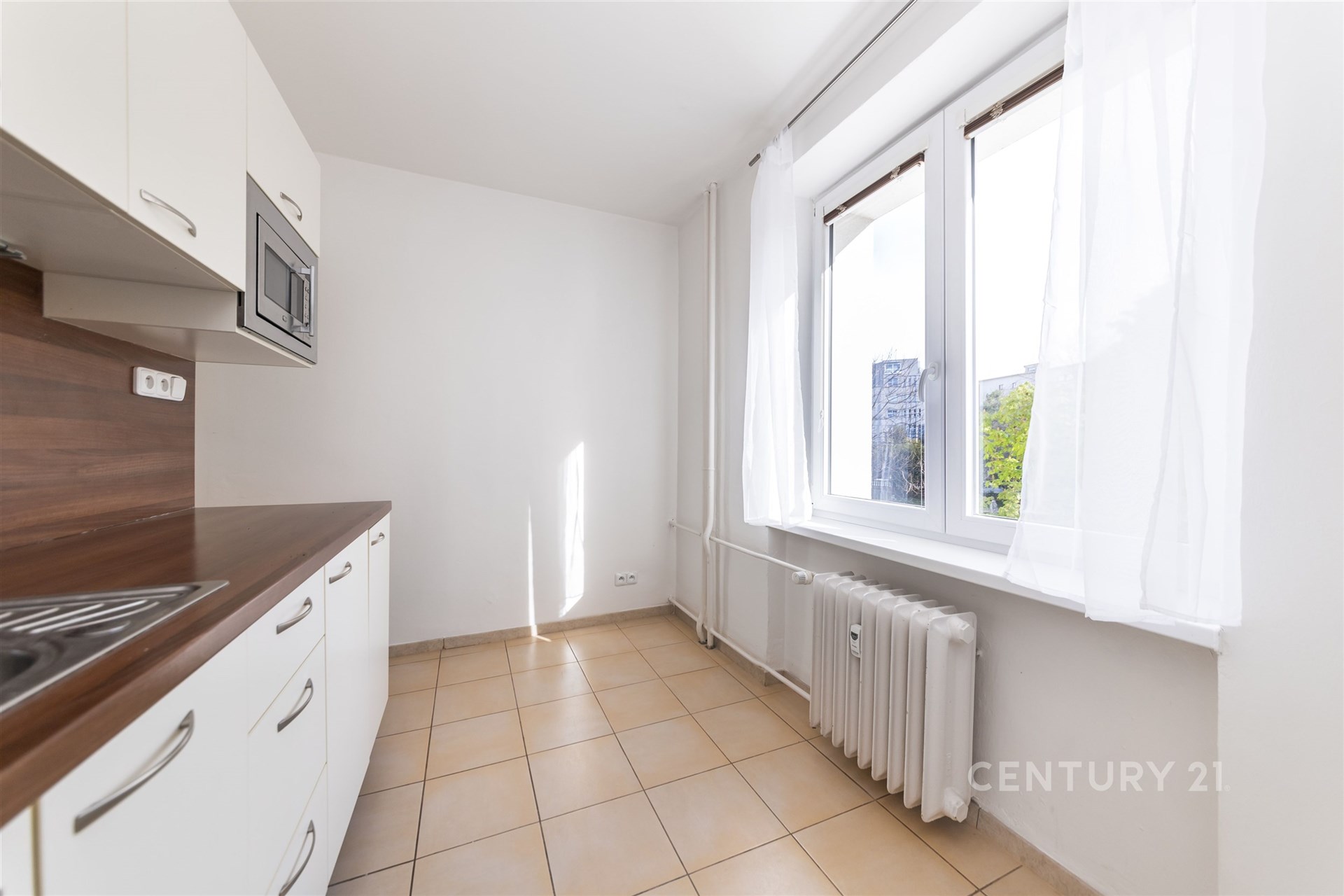 Světlá kuchyň s dlažbou, vestavěné skříně, velká okna, moderní design, bílé stěny vytvářejí prostorný a příjemný prostor.