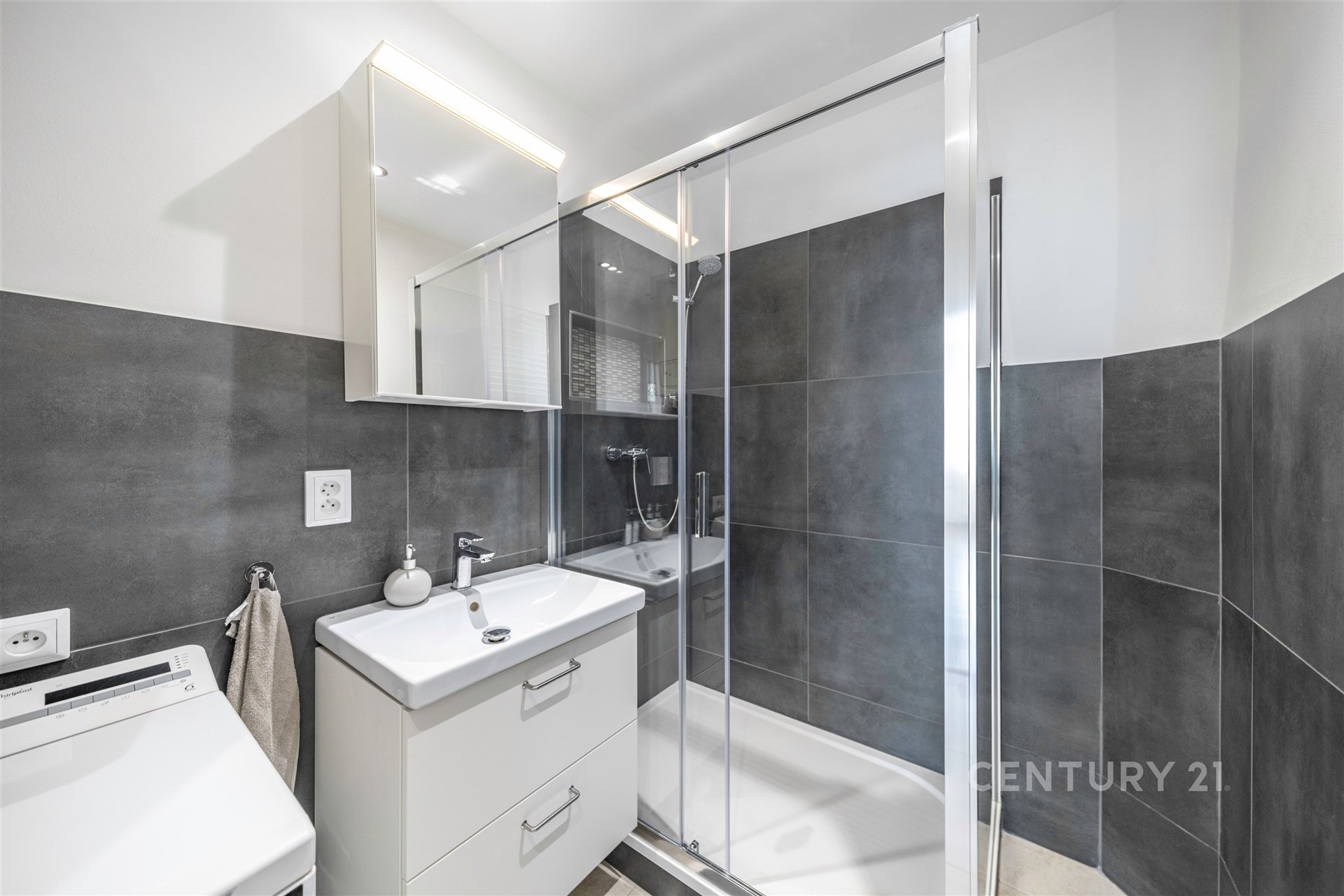 Moderní koupelna s prostorným sprchovým koutem, šedými obklady a elegantním nábytkem. Velké zrcadlo a čistý design vytvářejí světlý prostor.